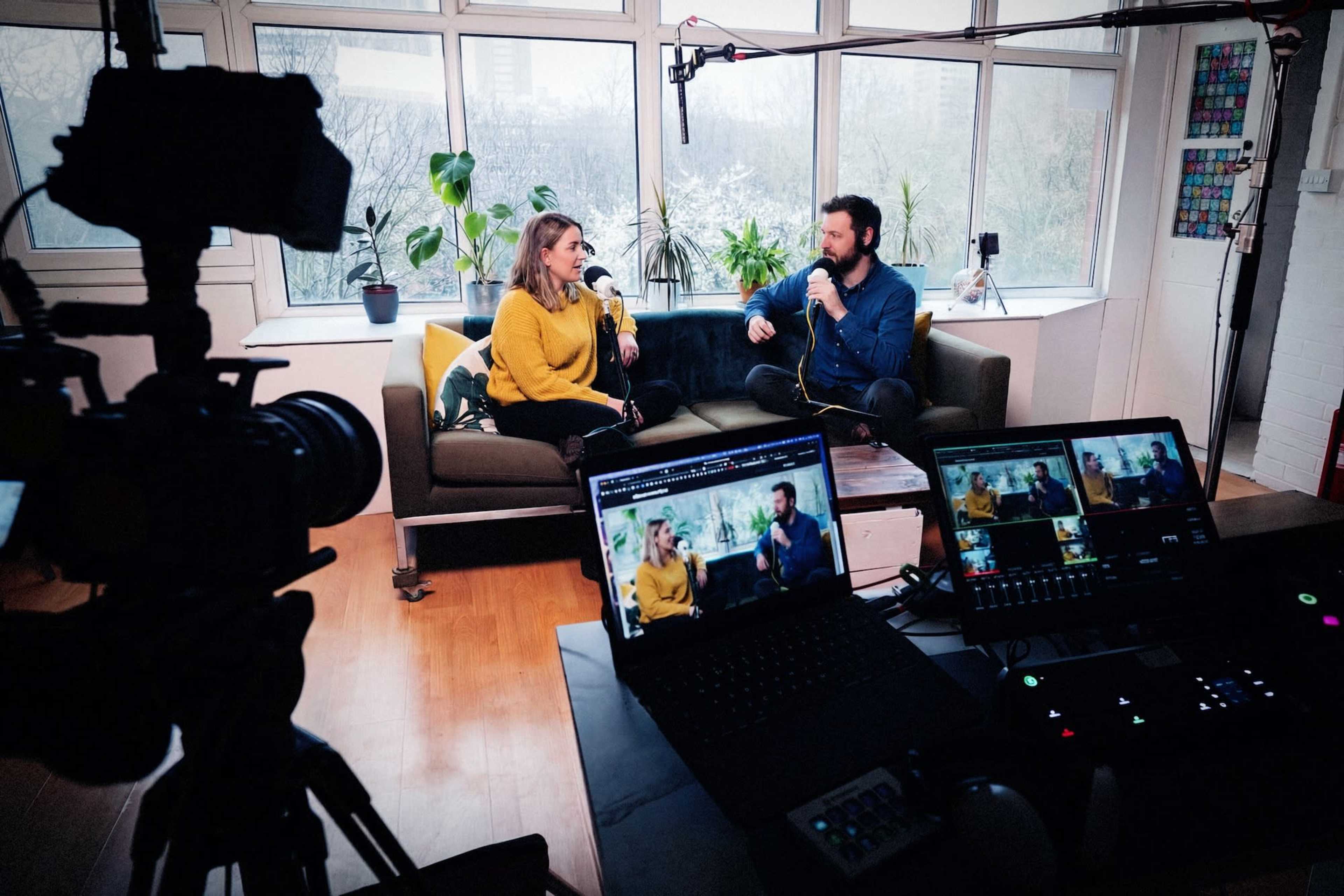 Two people are seated on a couch in a well-lit room, engaging in a discussion while microphones are positioned nearby and filming equipment is set up in the foreground.
