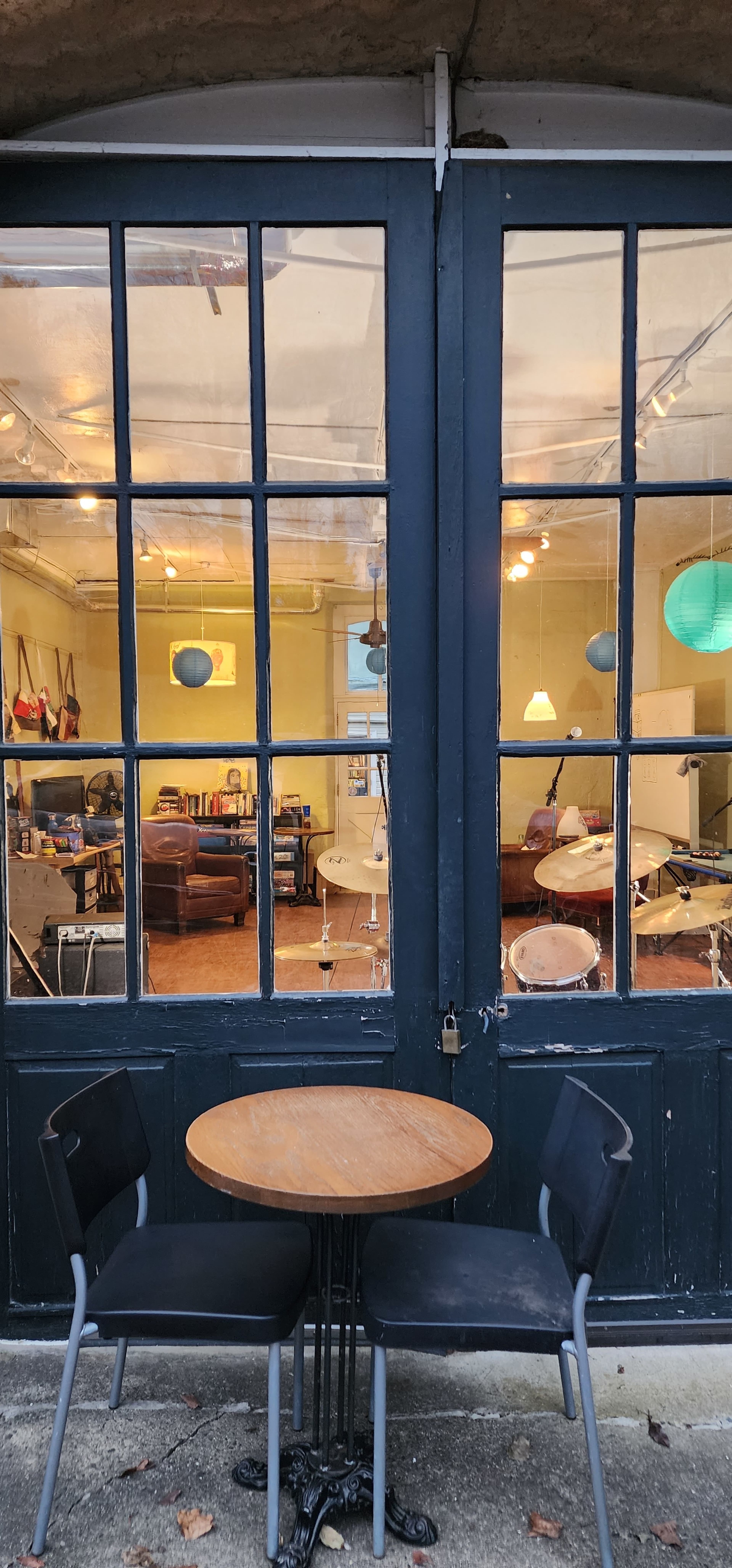 A round wooden table with two black chairs is positioned outside a glass door that leads into a lit room furnished with sofas and hanging light fixtures.