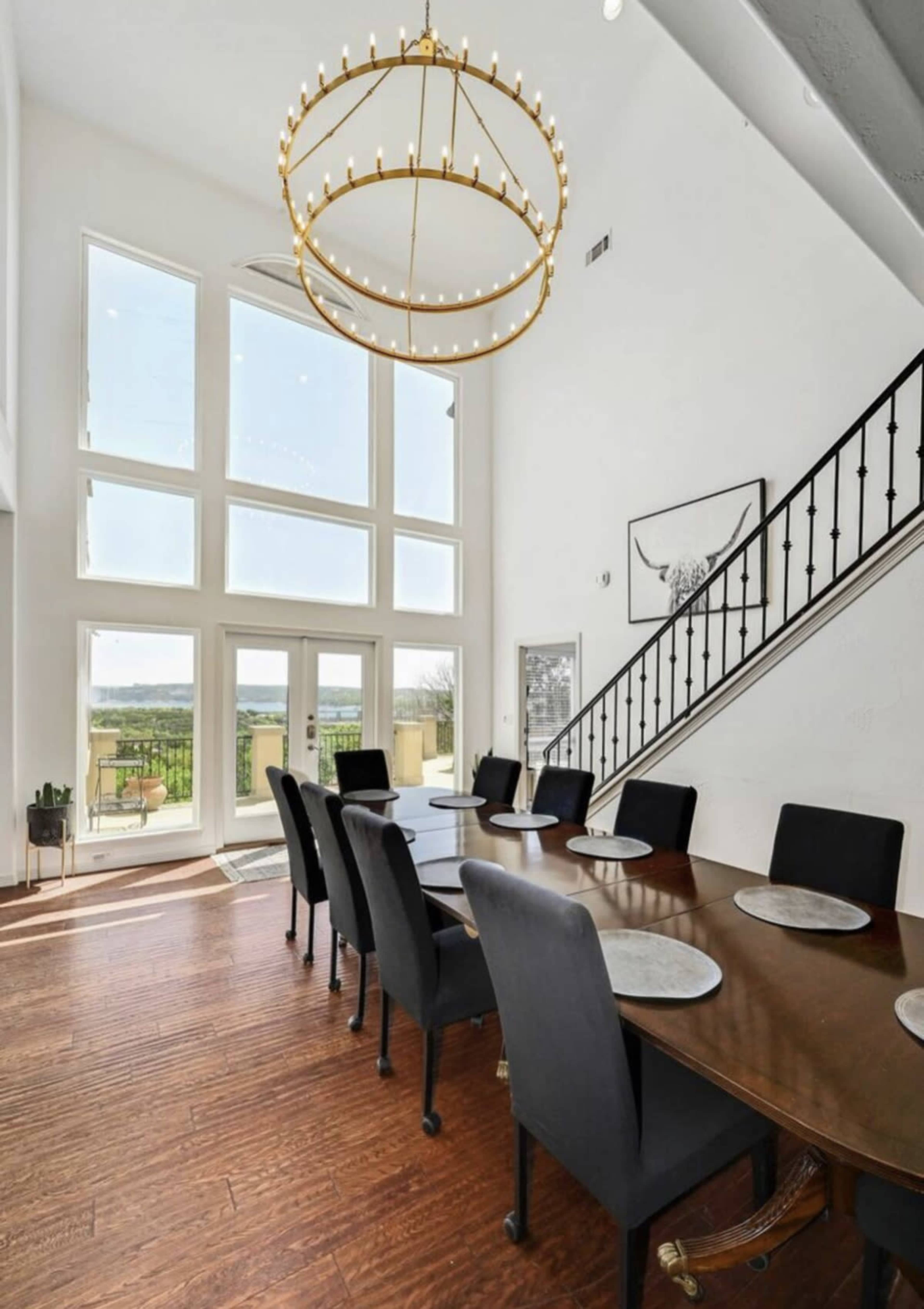 A dining room with a large wooden table set for eight, surrounded by black chairs, and featuring tall windows that allow natural light to illuminate the space.