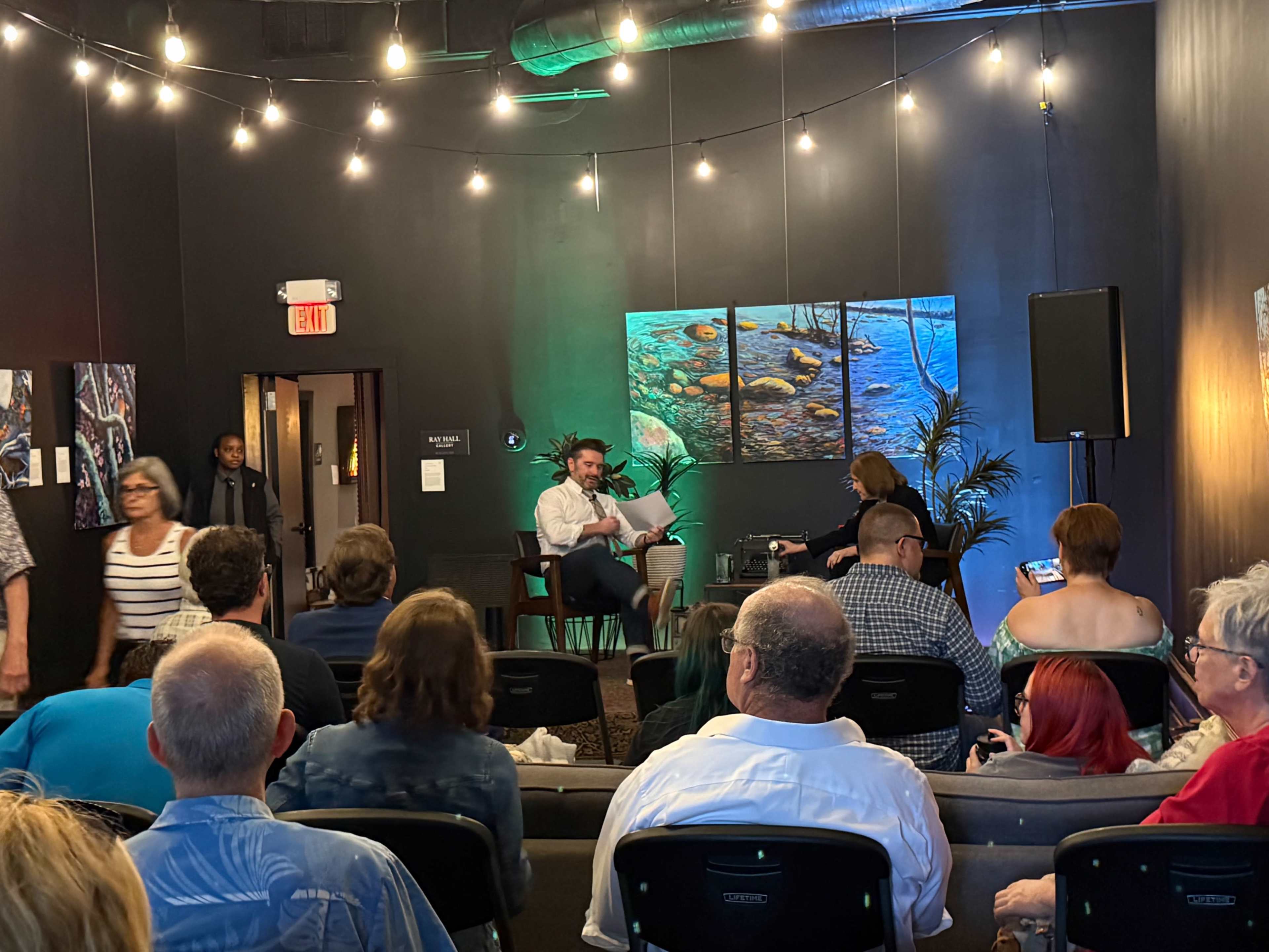 A small audience is seated in a dimly lit room with string lights, listening to a speaker on stage while a visual art piece is displayed on the wall.