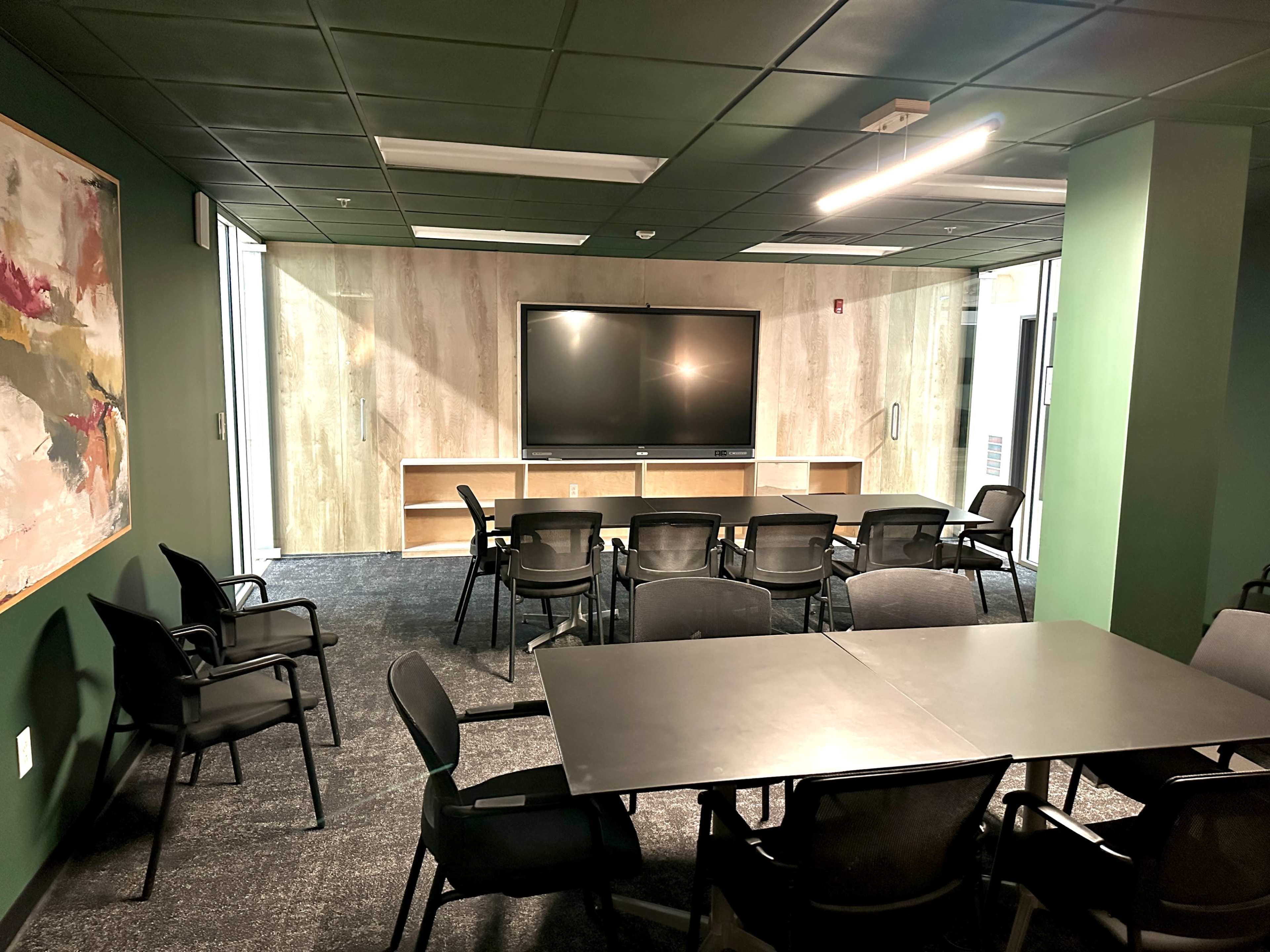 The image shows a modern conference room with a large television on a wall, surrounded by several black chairs and tables, all set against a green and wooden backdrop.