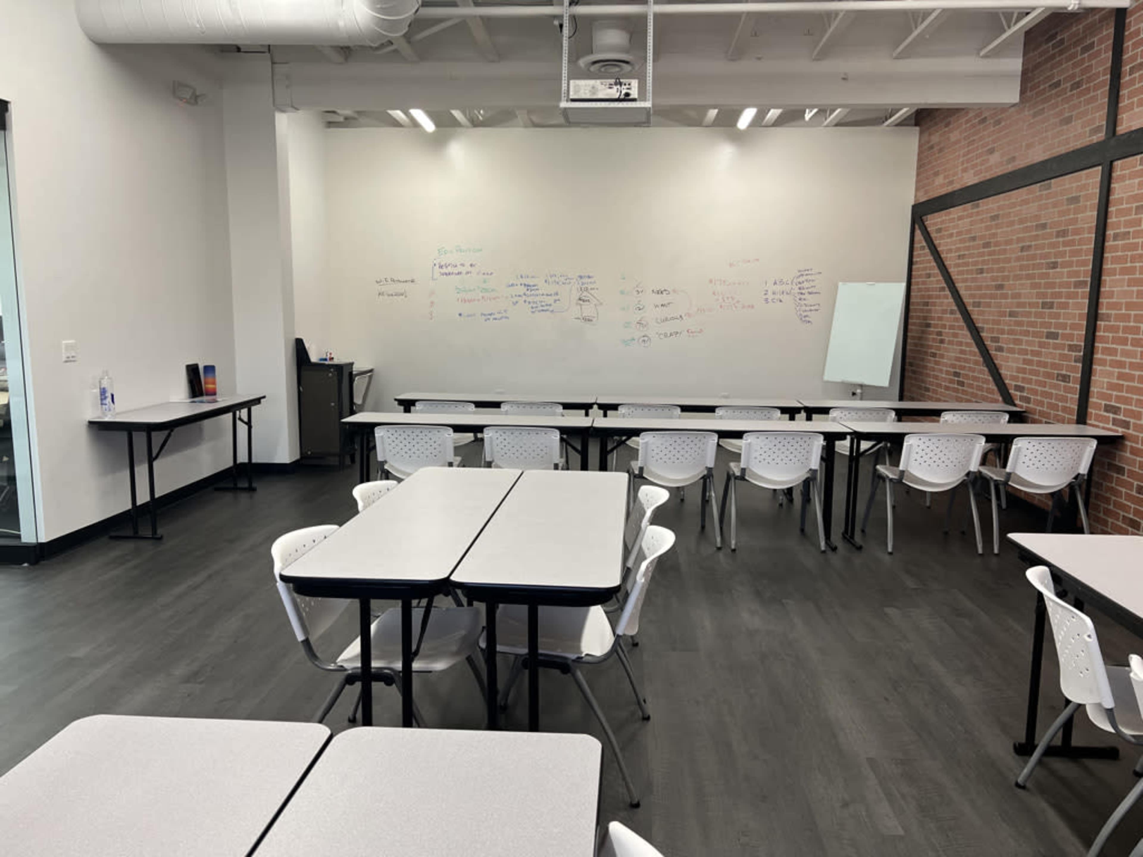 A classroom with several tables and chairs arranged around the room, a projector on the ceiling, and whiteboards on one wall.