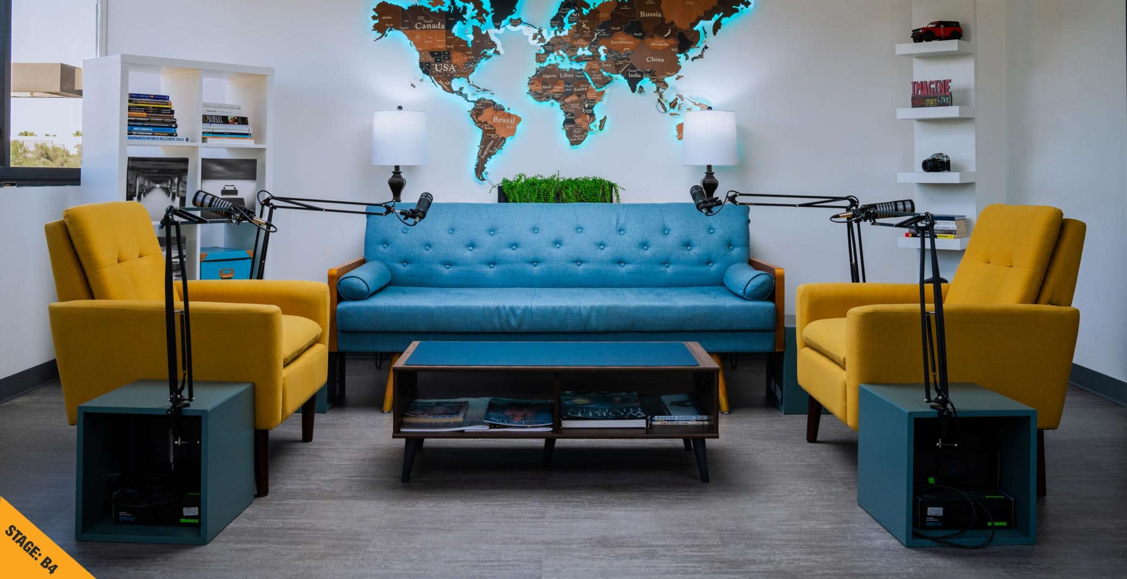 The scene features a modern lounge area with a blue couch, two yellow chairs, a coffee table, and microphones positioned for recording.