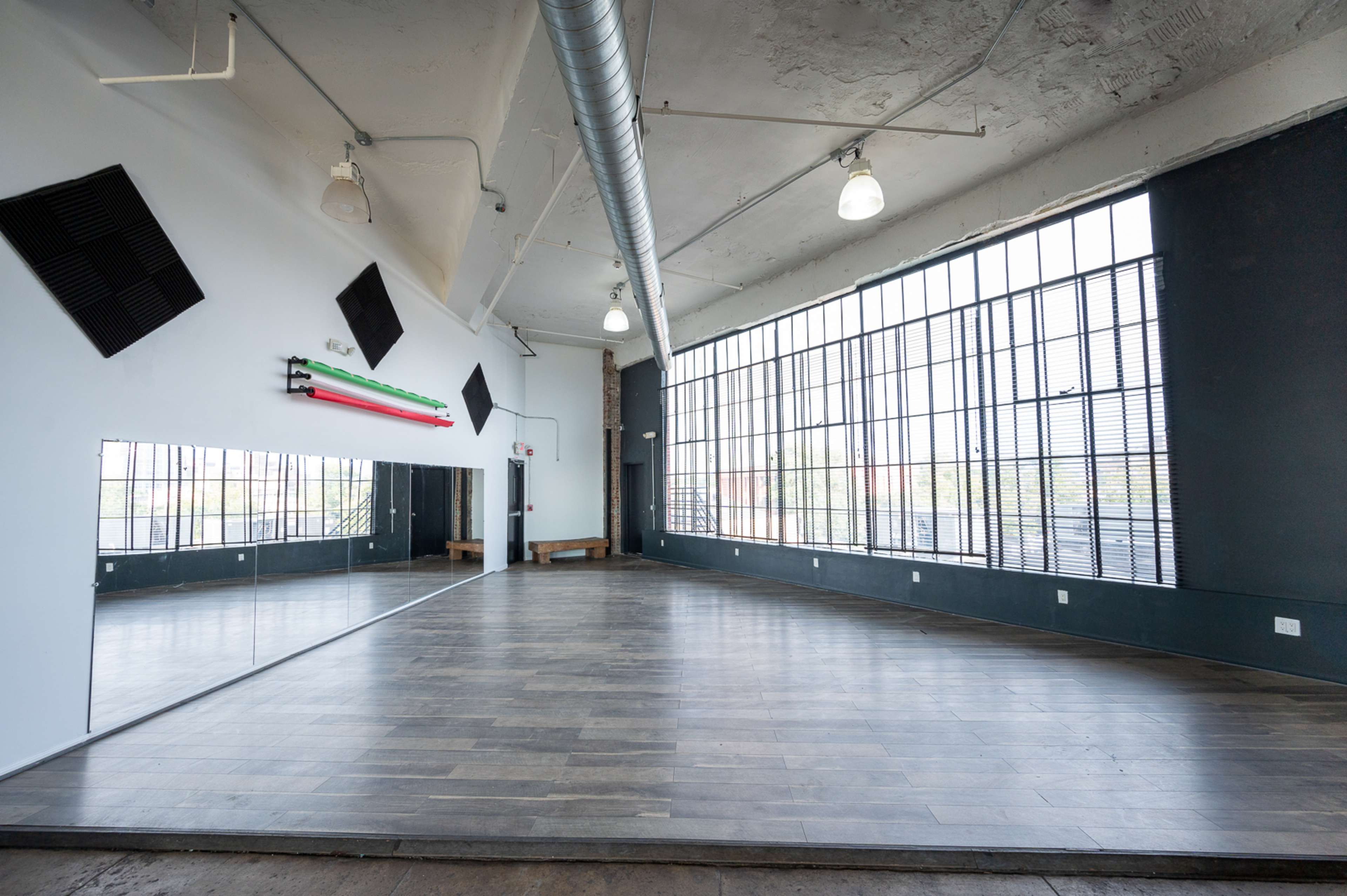 A spacious, well-lit room features large windows, wooden flooring, and a mirror along one wall.