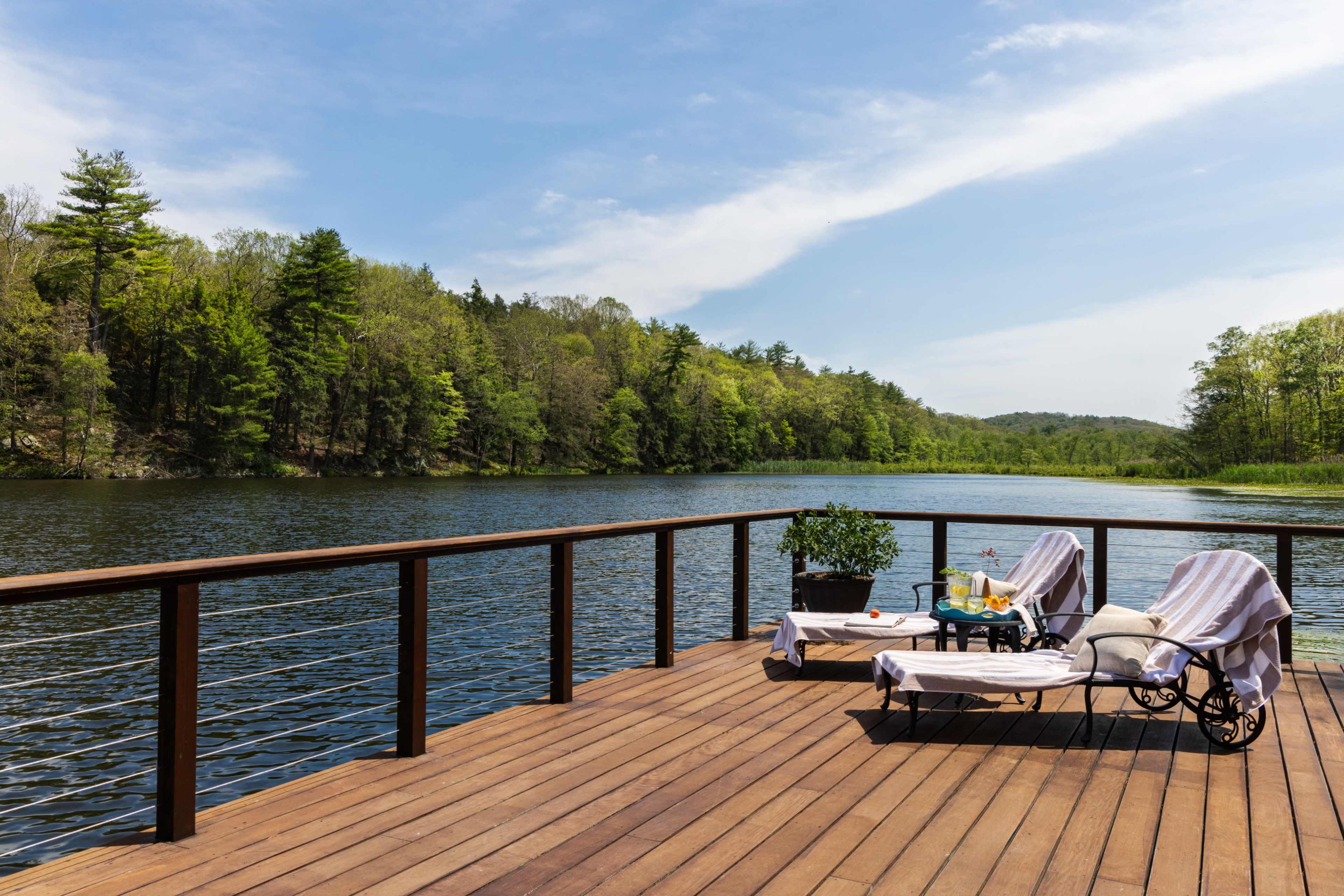 A wooden deck with two lounge chairs overlooks a calm lake surrounded by lush greenery.