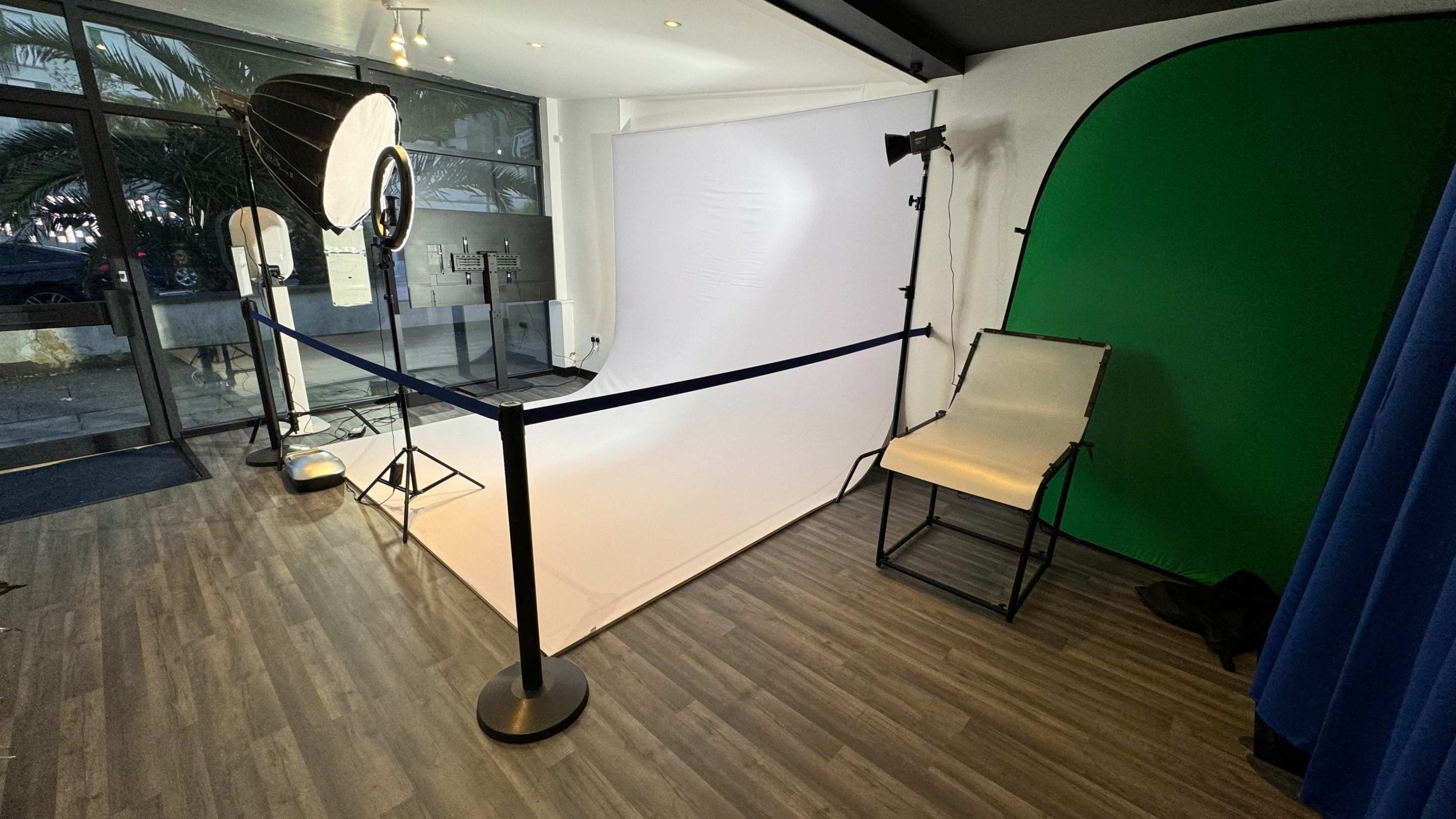 A photography studio setup with a white backdrop, a green screen, lighting equipment, and a folding chair.
