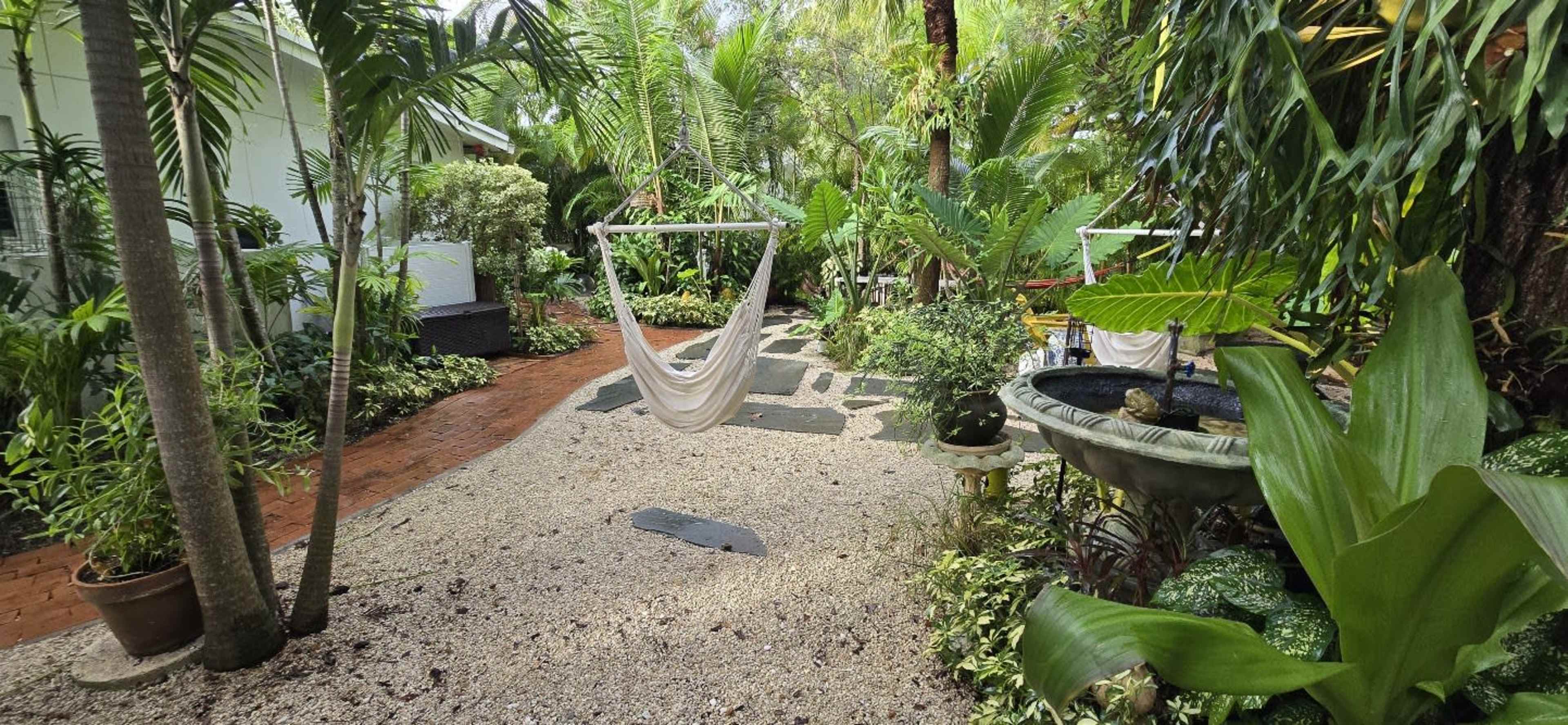 A serene garden pathway is lined with tropical plants, a hammock hangs between two trees, and there is a stone fountain surrounded by lush greenery.