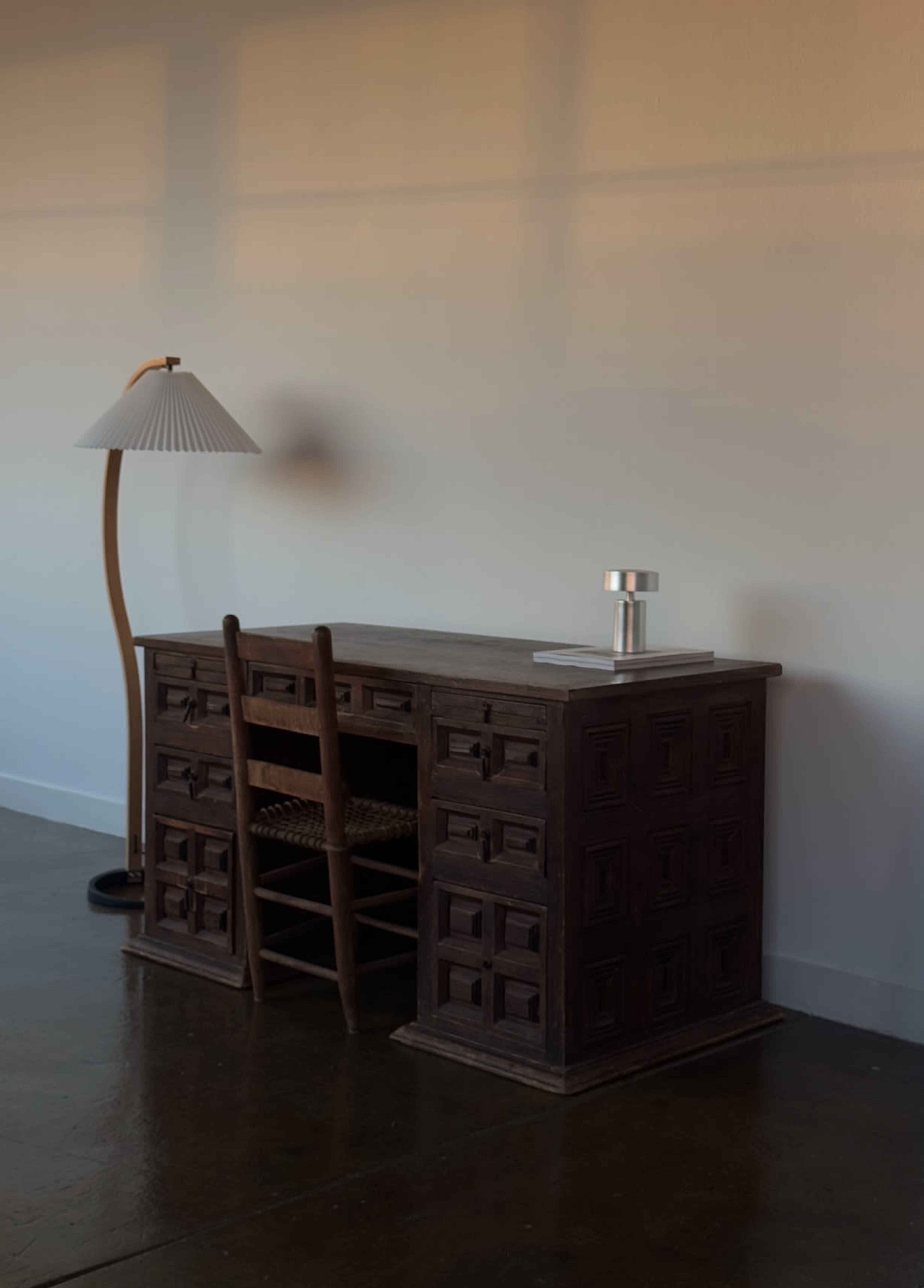 The image shows a wooden desk with multiple drawers, accompanied by a chair and a standing lamp, set against a plain wall.