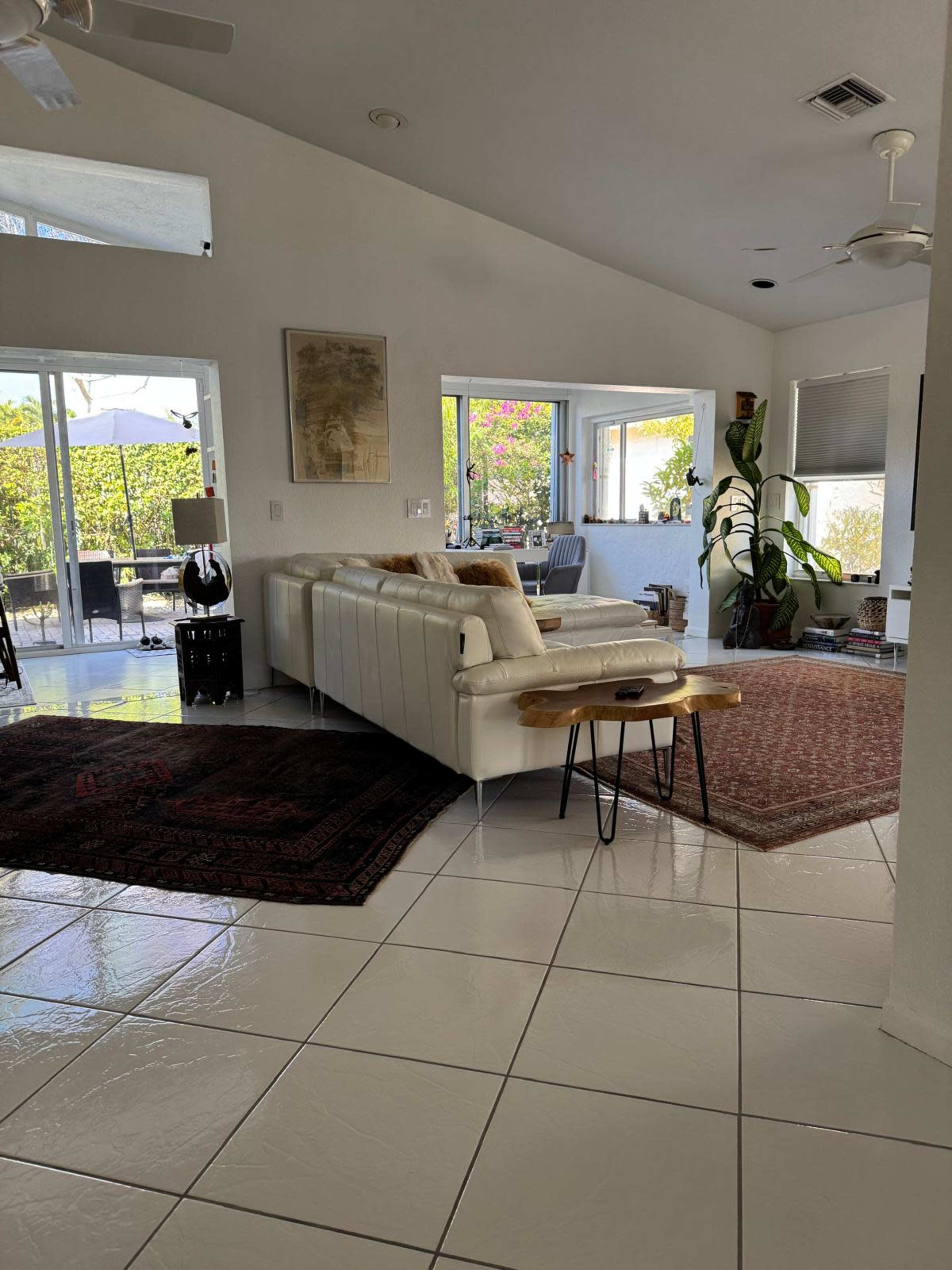The image shows a spacious living room with a white sectional sofa, a large window overlooking a patio, and various decorative elements including a rug and indoor plants.