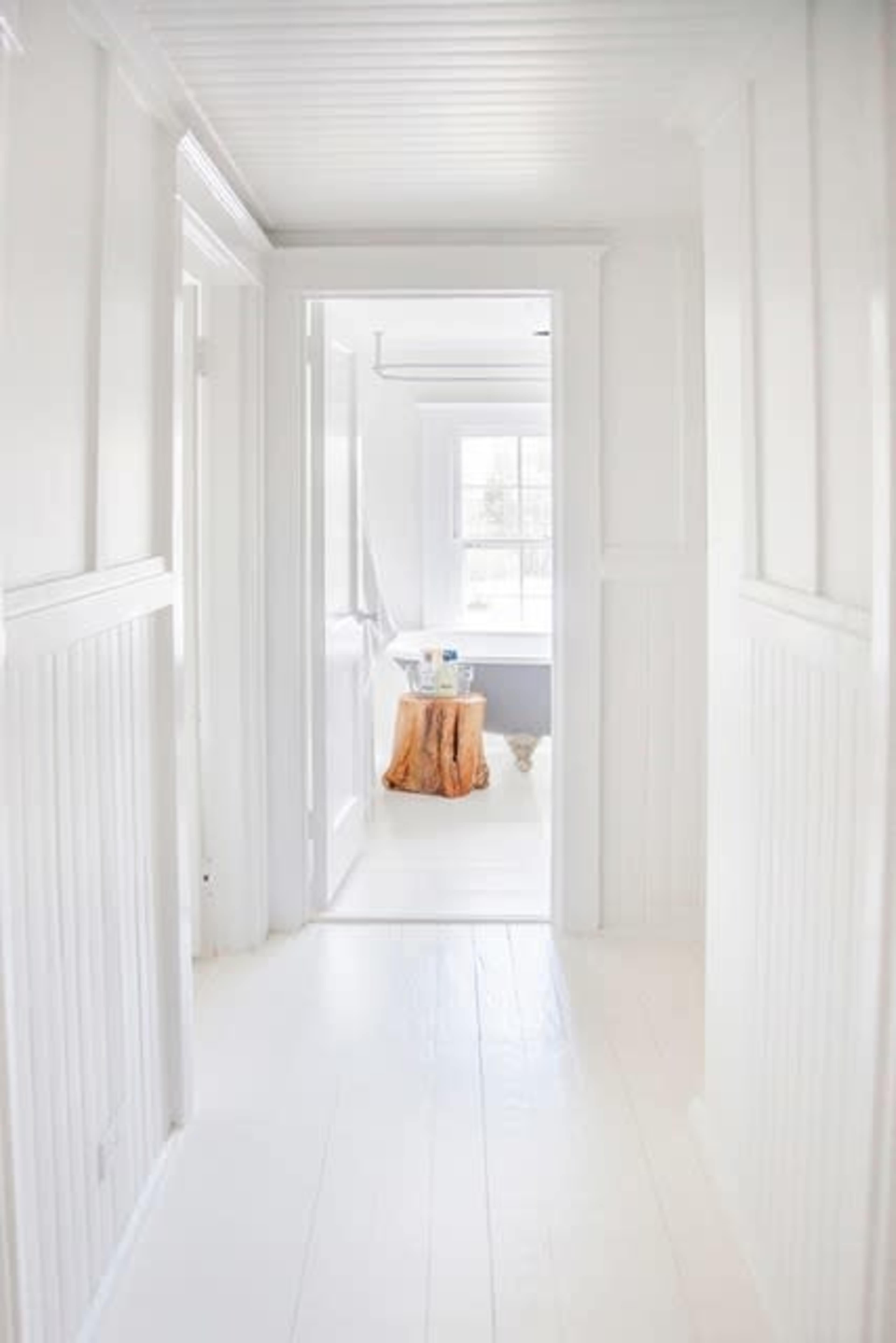 A bright, white hallway leads to a room with natural light and a wooden side table.