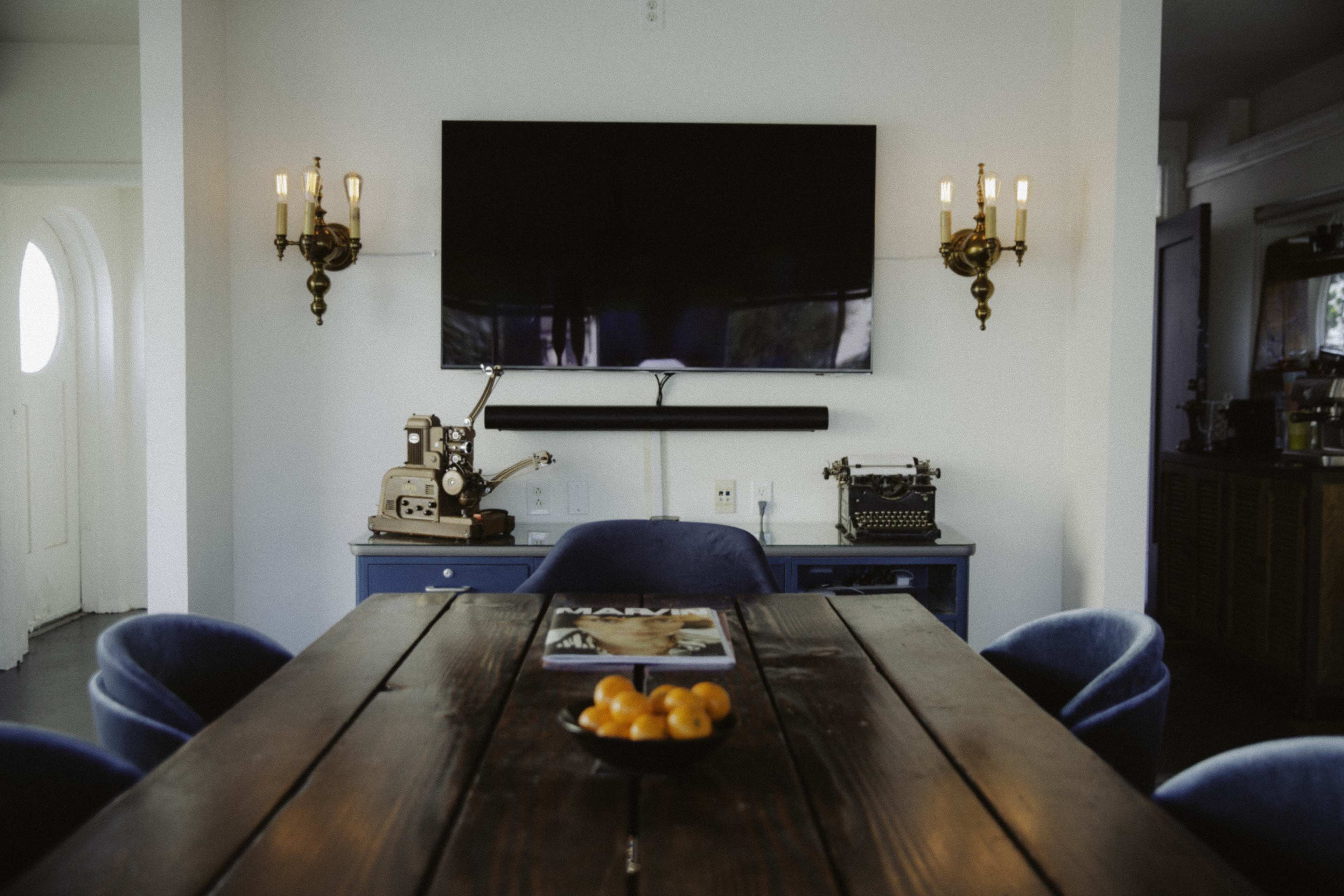 A rectangular wooden table is centered in a room with a large television mounted on the wall, flanked by wall sconces, and a vintage typewriter displayed on a blue cabinet.