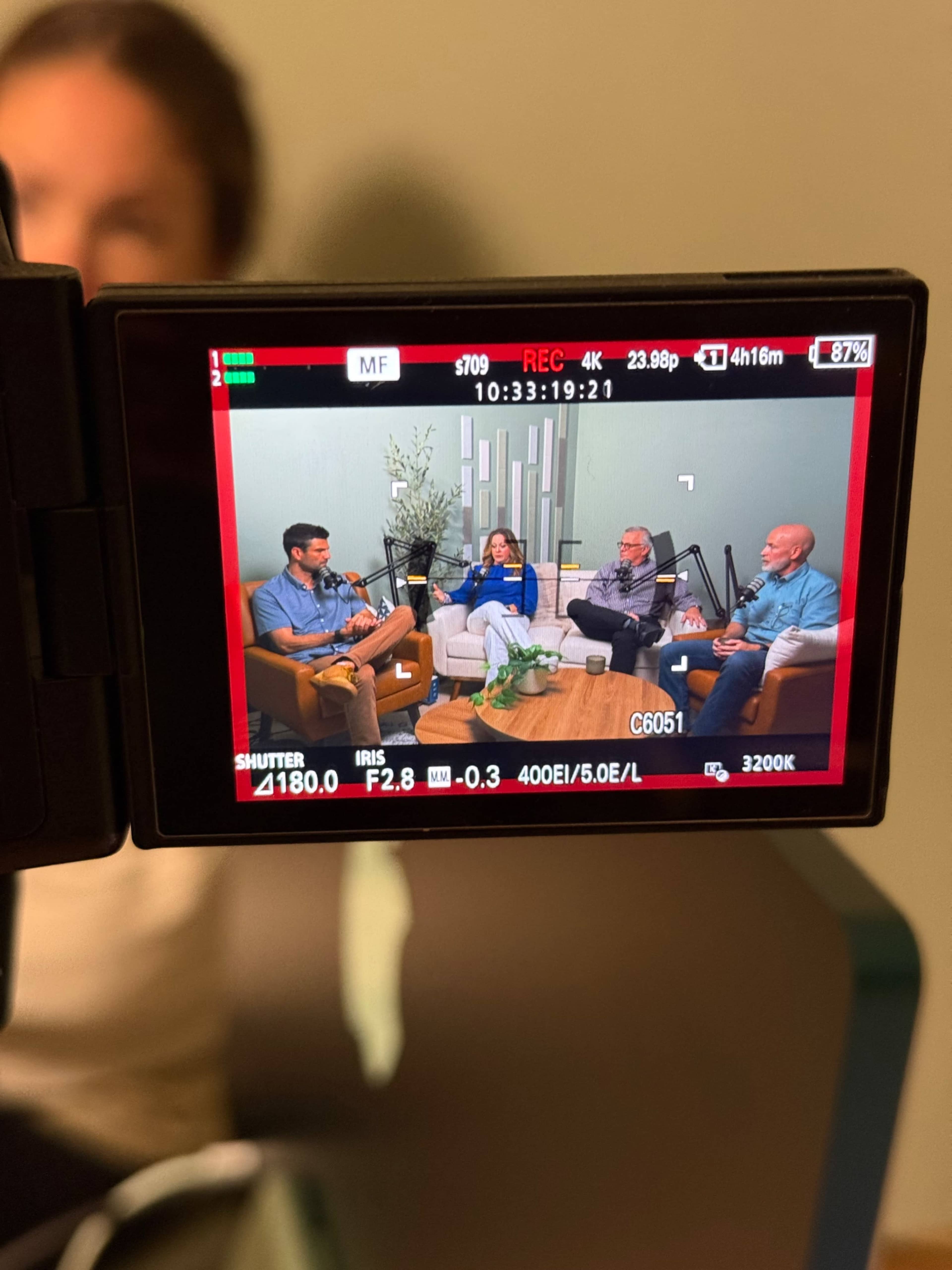 A video recording displays four individuals seated in a casual setting, engaged in conversation with decorative plants in the background.