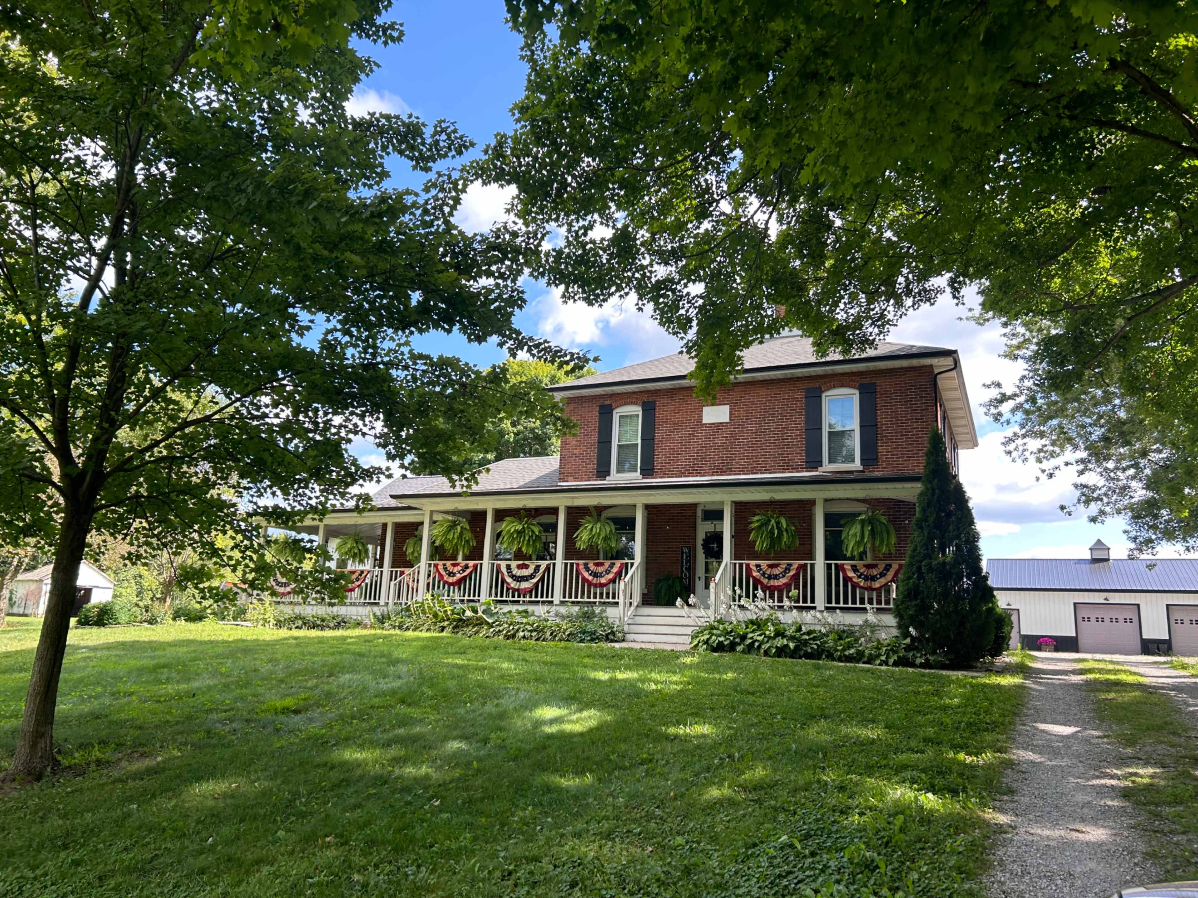 A two-story brick house with a covered porch is surrounded by trees and greenery, featuring patriotic decorations.
