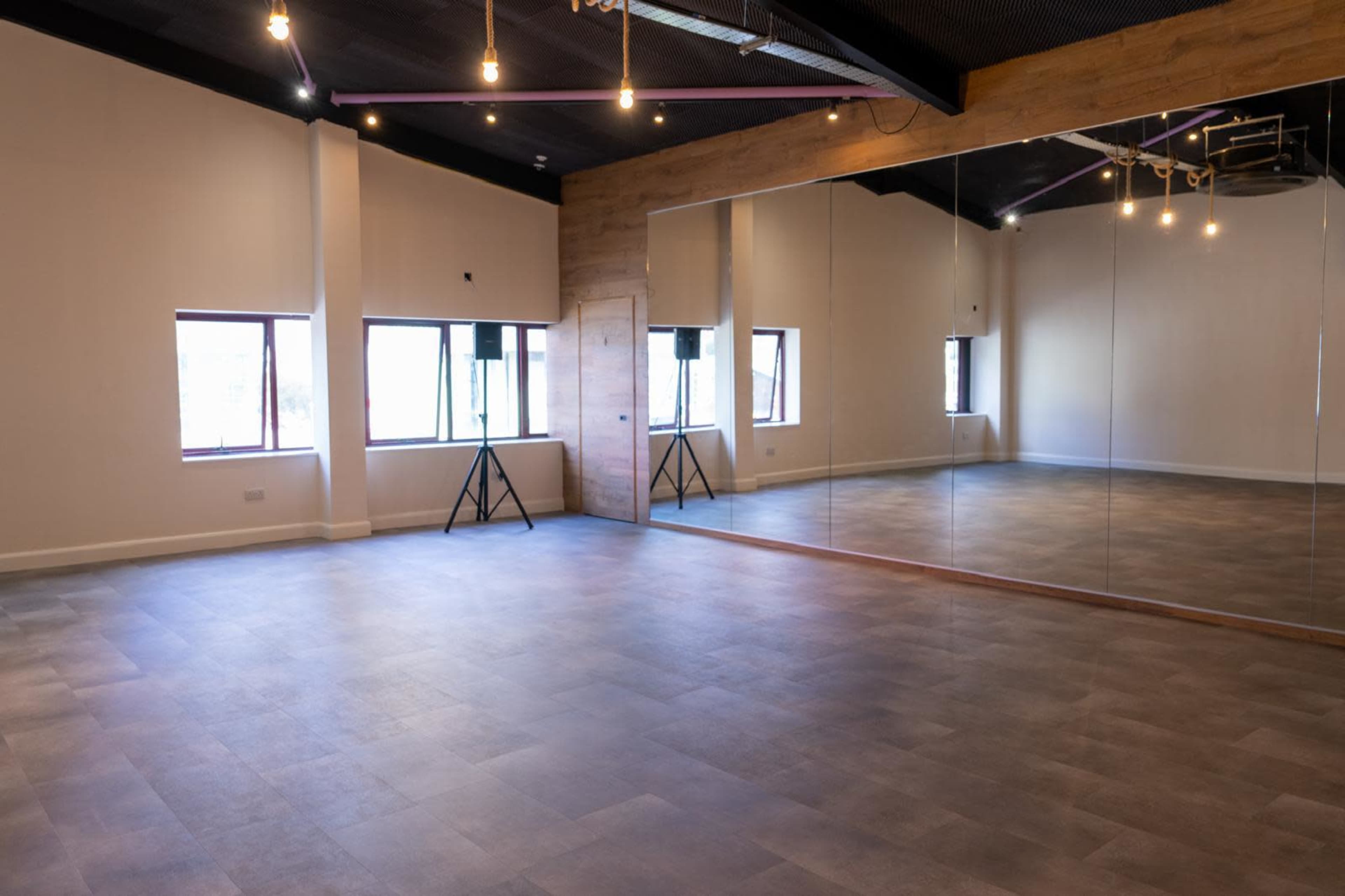 The image shows a spacious, light-colored room with a mirrored wall, two speakers on stands, and natural light coming through the windows.