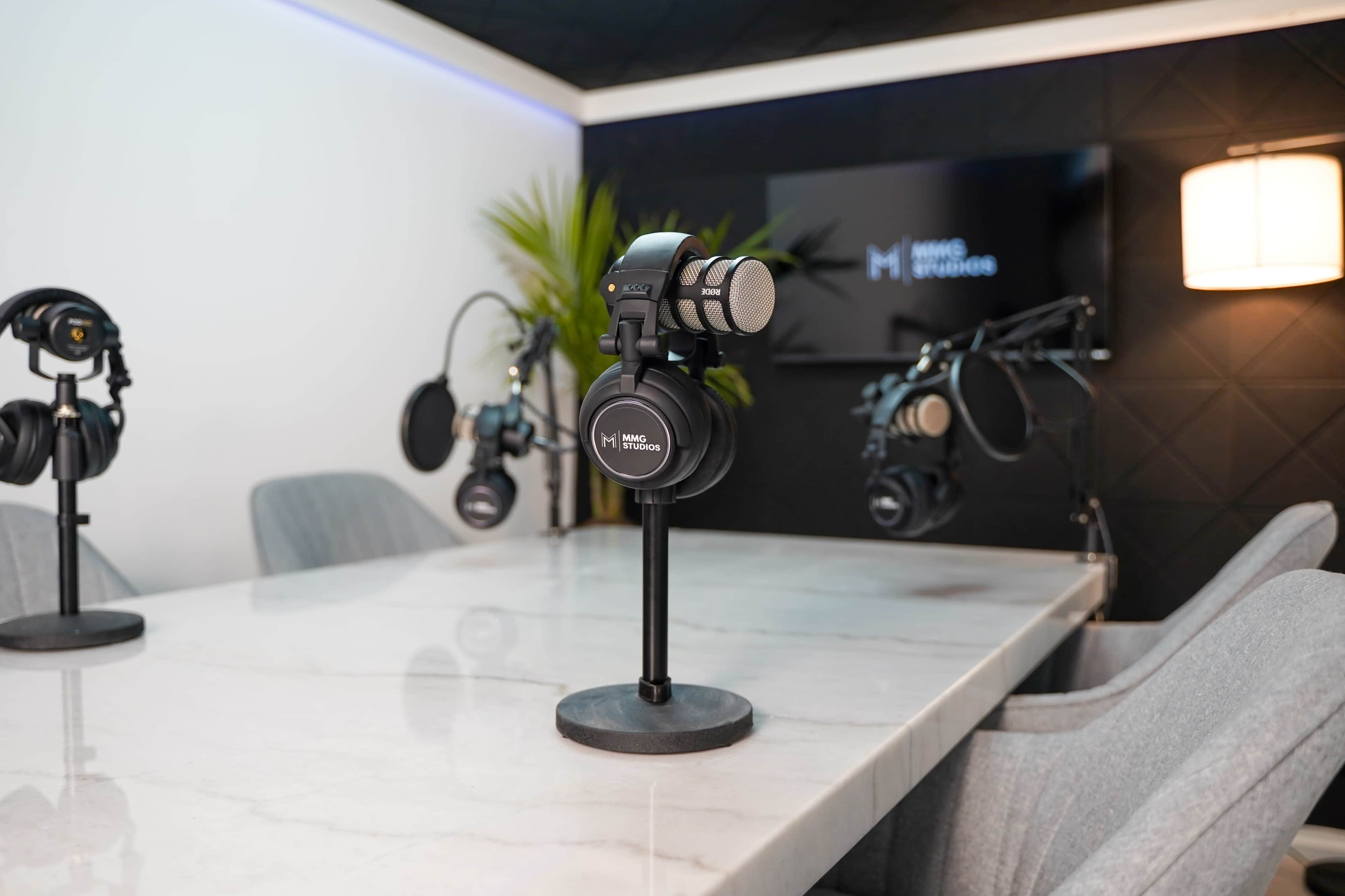 A modern podcast studio features multiple microphones on stands, headphones, and a sleek marble table against a dark wall with a television.