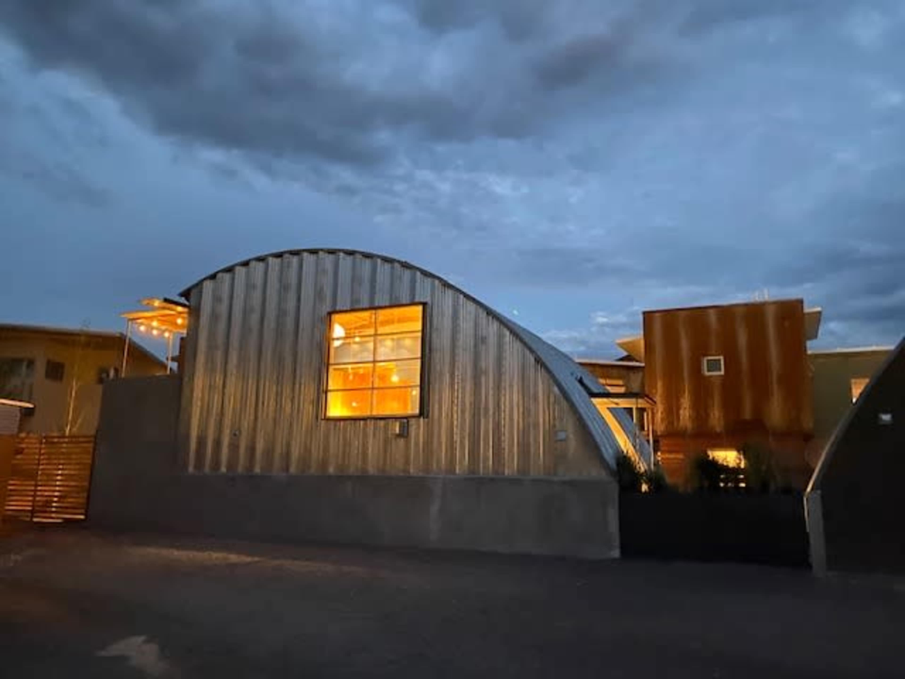 A metal-sided building with a curved roof and glowing windows stands illuminated under a cloudy twilight sky.