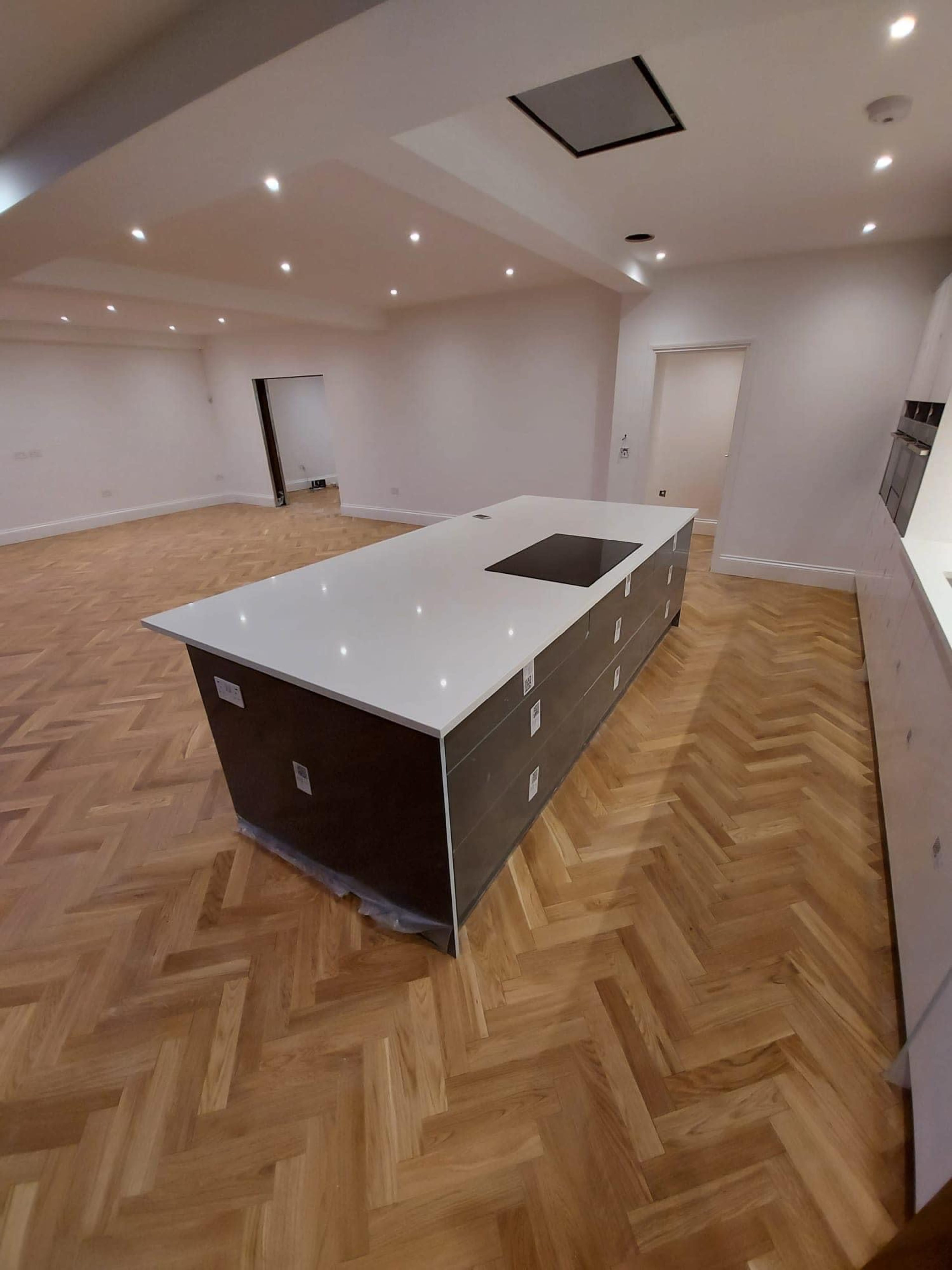 A spacious kitchen with a central island featuring a white countertop and dark cabinetry, surrounded by herringbone-patterned wooden flooring and recessed lighting.