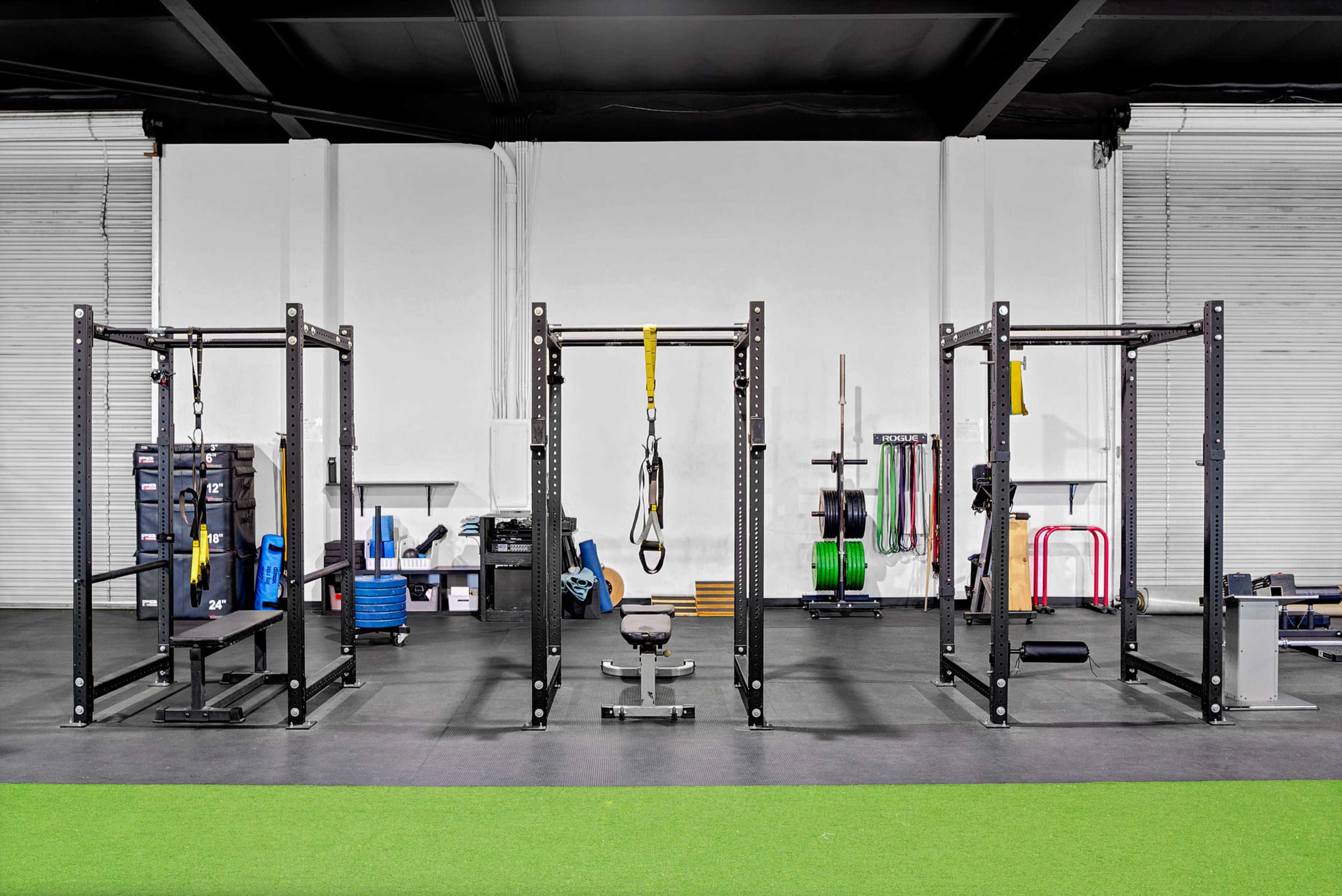 A spacious gym area with multiple weightlifting racks, a bench, and exercise equipment arranged on a black floor with a green turf section.