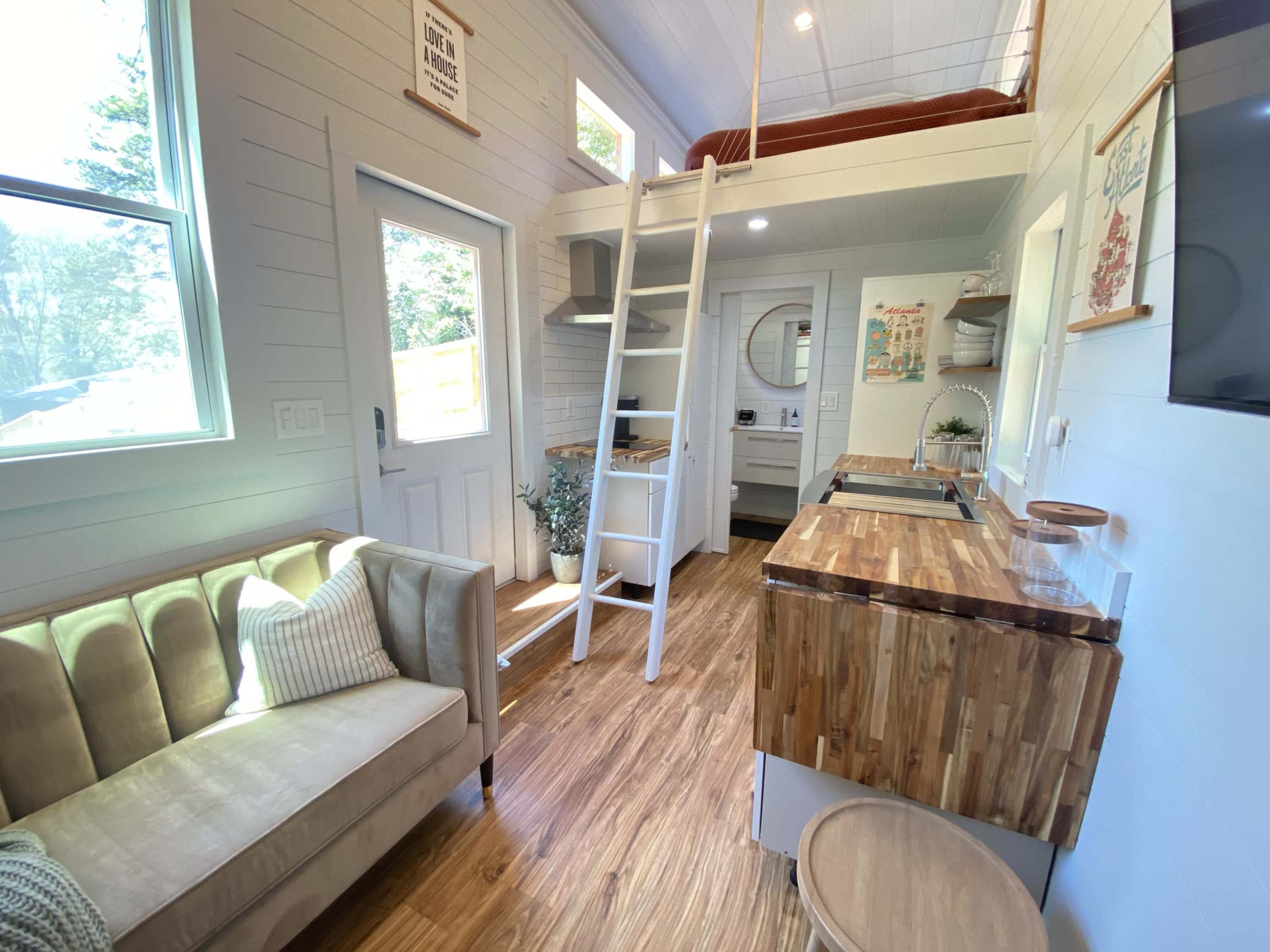 The image shows a cozy interior of a tiny house featuring a ladder to a lofted bed, a kitchenette with wooden countertops, and a living area with a beige sofa.