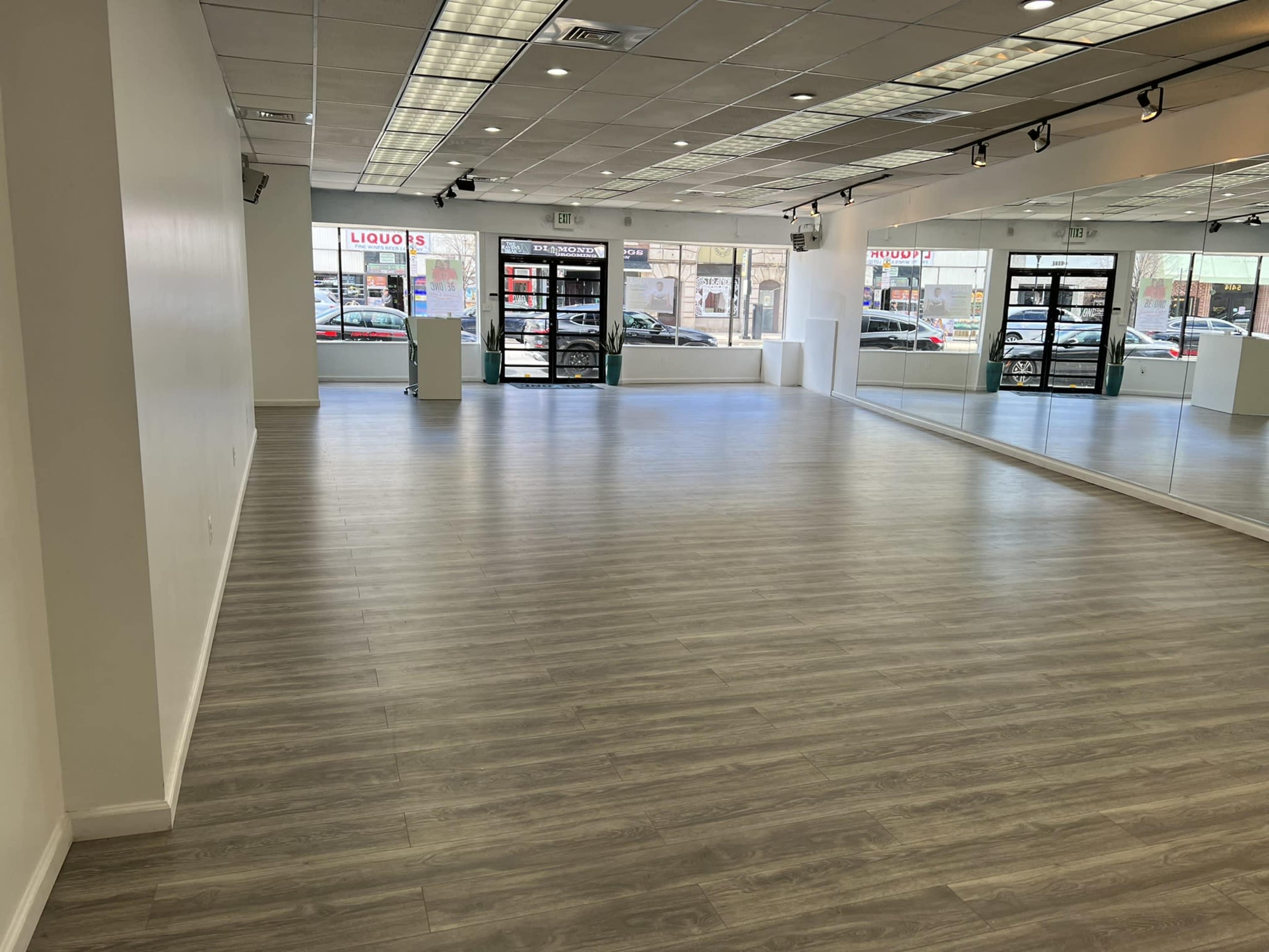 A spacious, empty room with light wood flooring, large mirrors on one wall, and windows facing the street.
