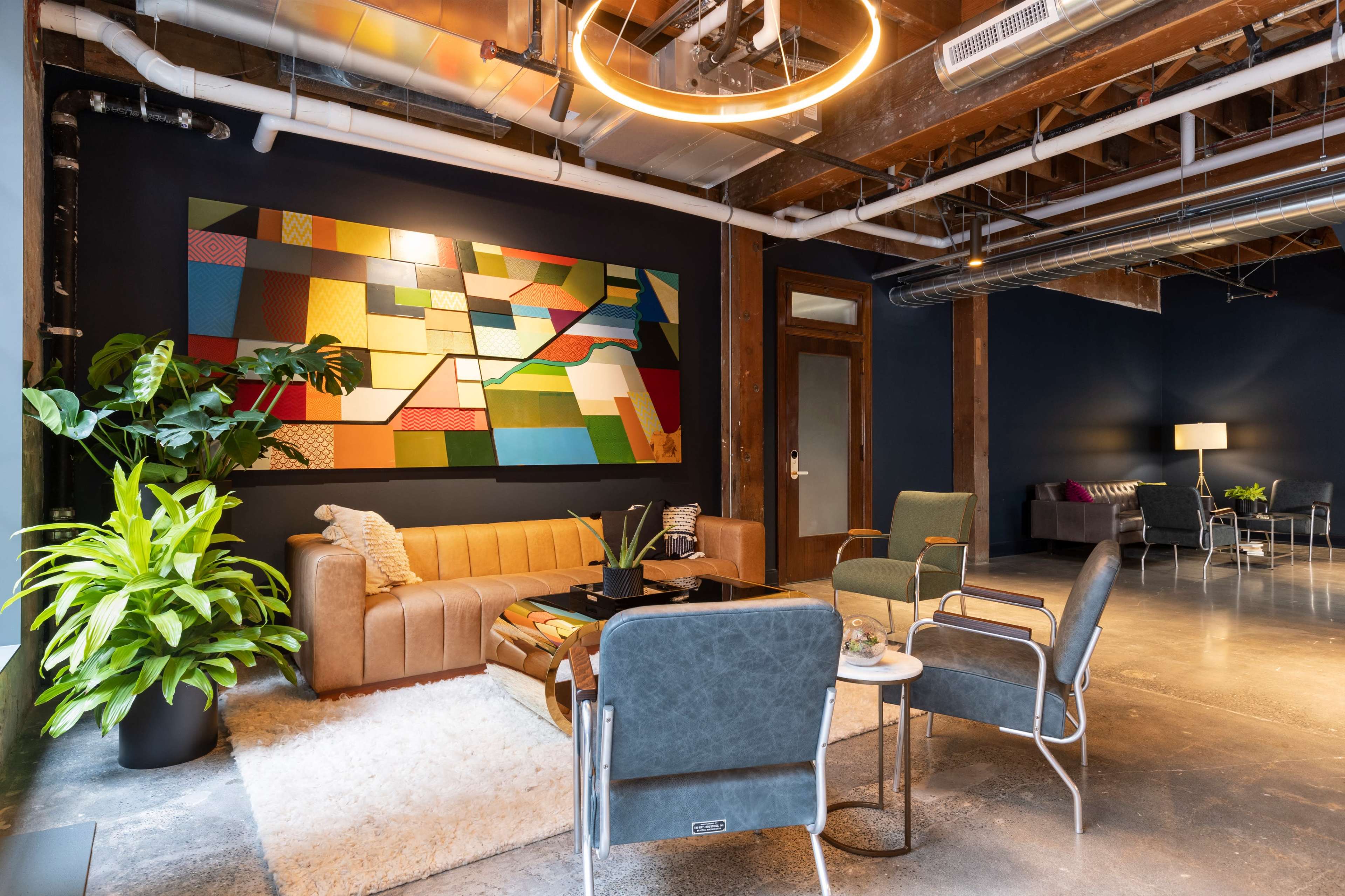 A modern, multi-functional lounge area featuring a brown sofa, potted plants, a large abstract painting on the wall, and a mix of seating options including armchairs and small tables.