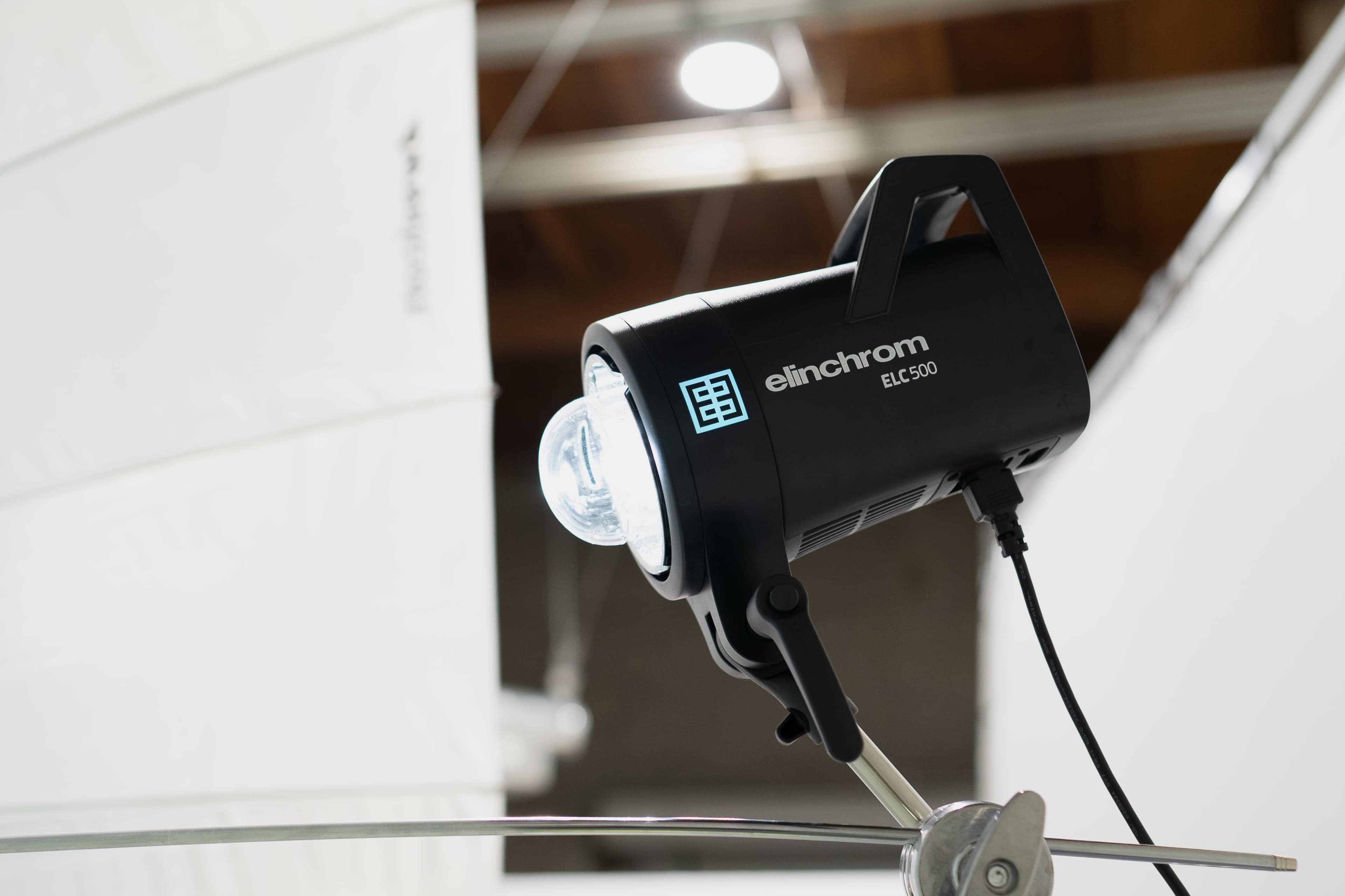 A black Elinchrom lighting unit is mounted on a stand, with a translucent umbrella diffusing light in the background.