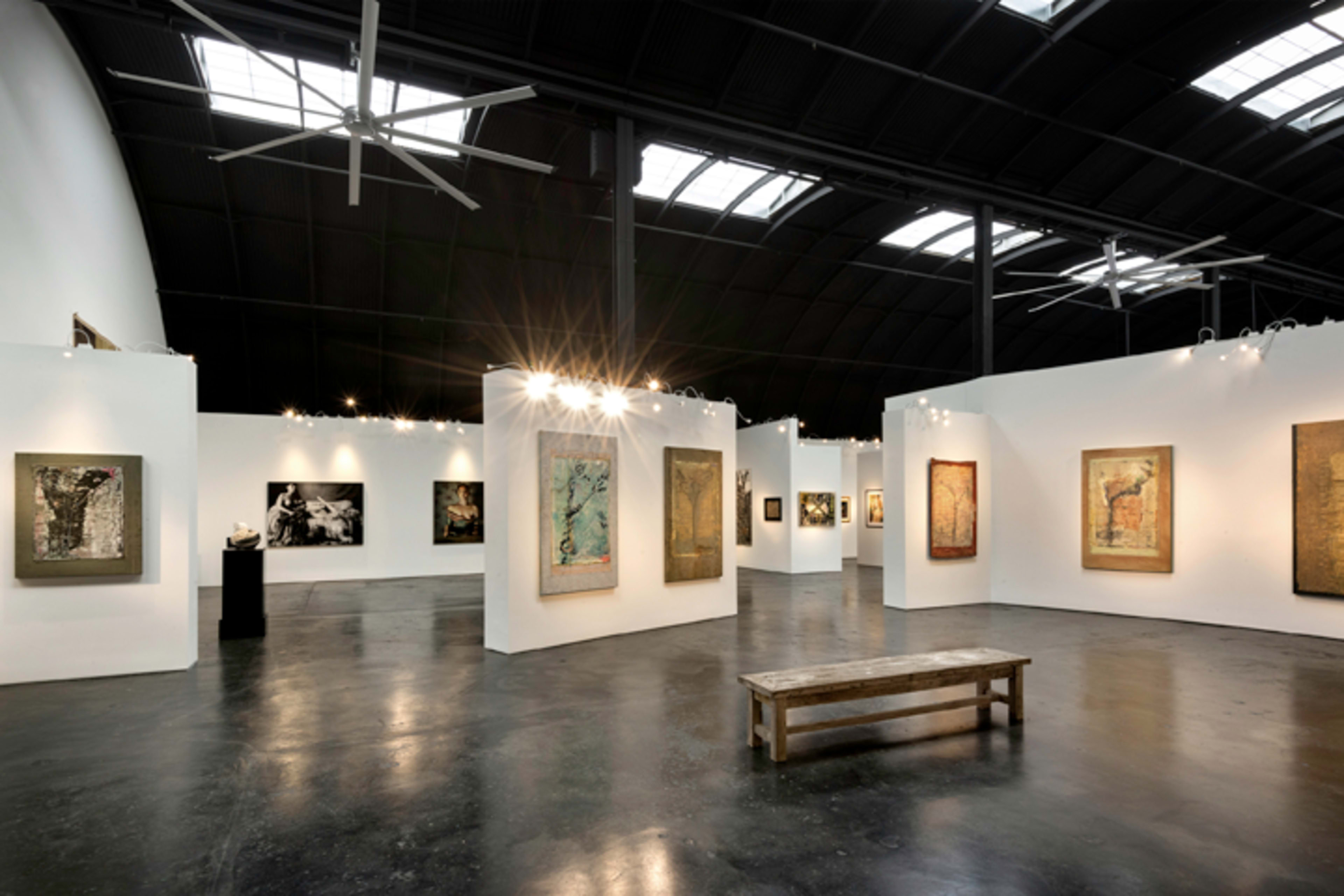 An art gallery with white walls displaying various framed artworks, and a wooden bench positioned in the center of the room.