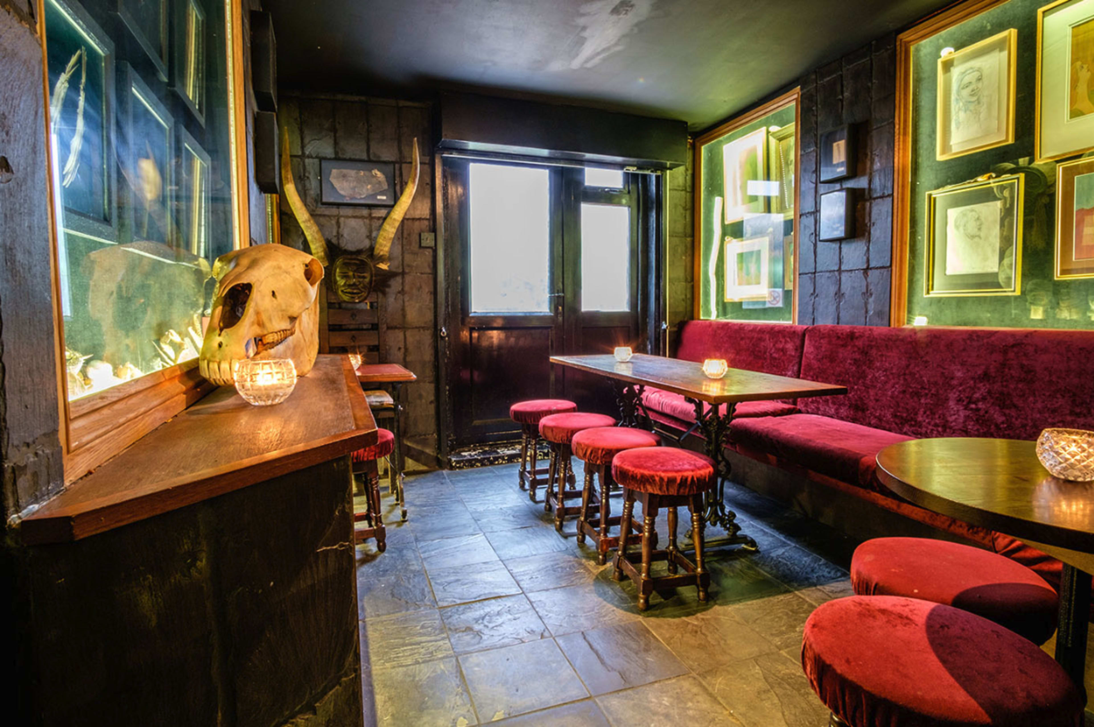 The interior of a dimly lit pub features red velvet seating, wooden tables, and decorative animal skulls on the walls.