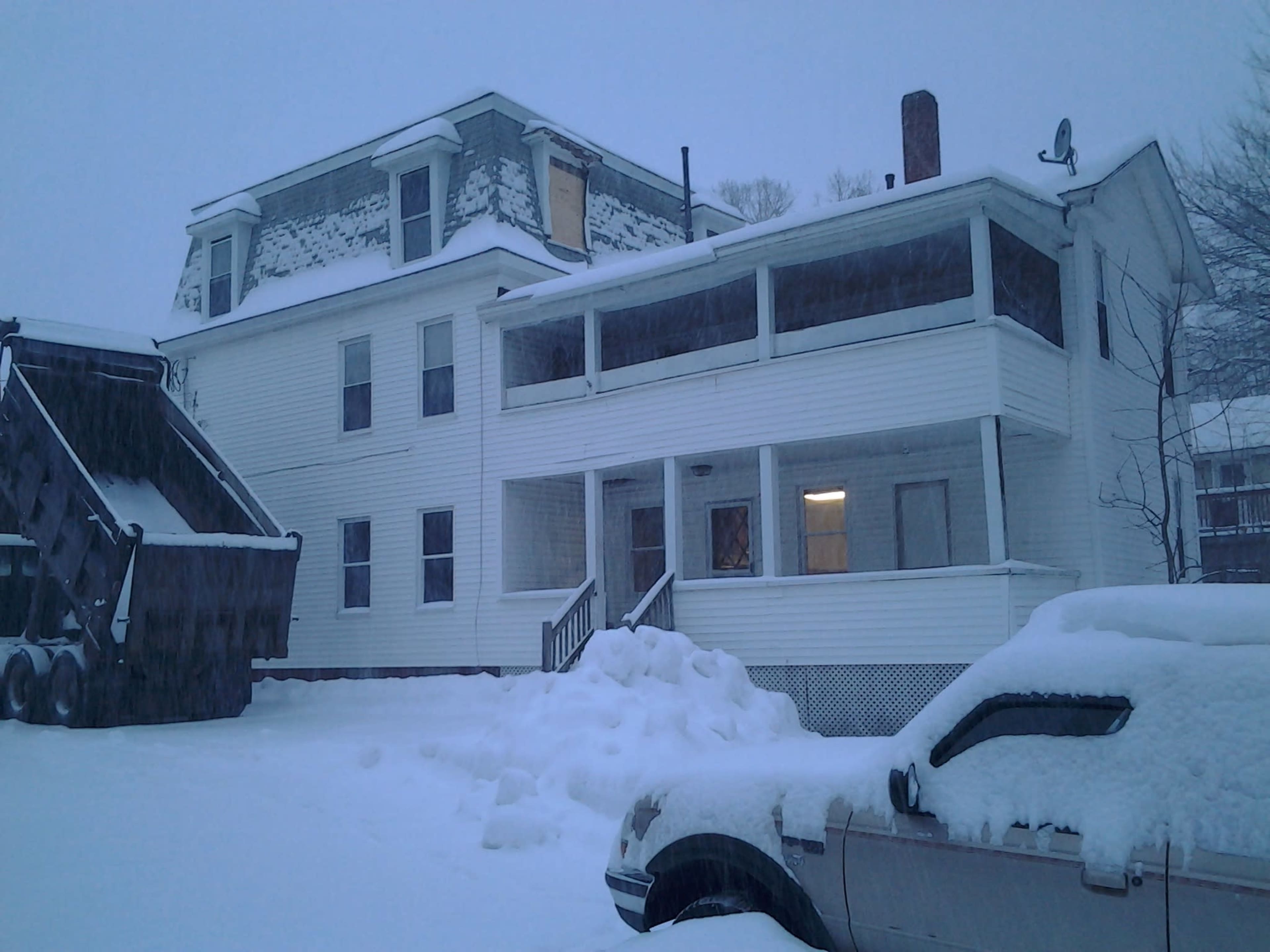 A white, multi-story building with a large porch is surrounded by several feet of snow, with a snow-covered vehicle parked nearby and a truck in the foreground.