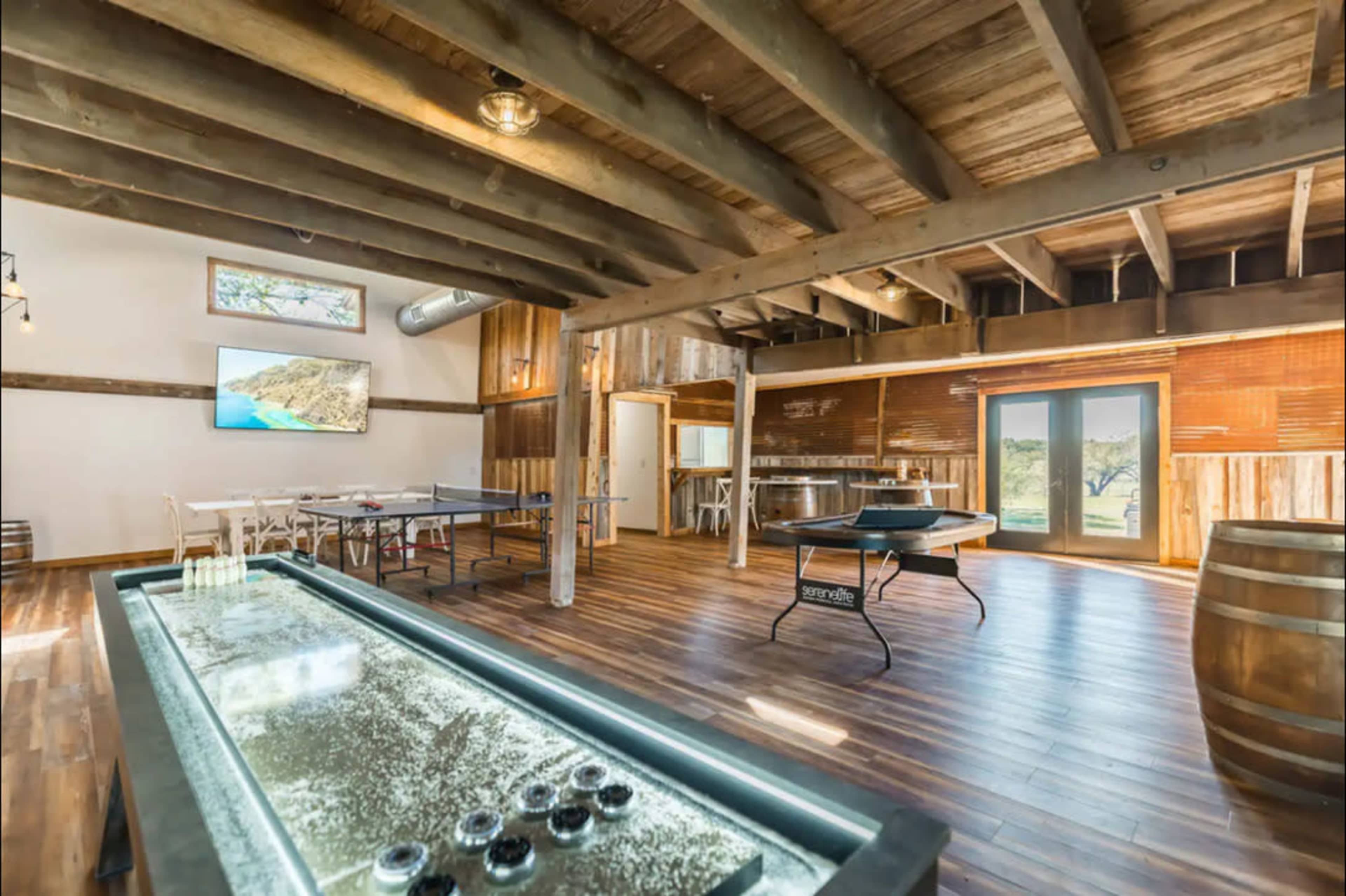 A spacious game room featuring a shuffleboard table, a ping pong table, and a large television on the wall, with wooden beams and a rustic design.