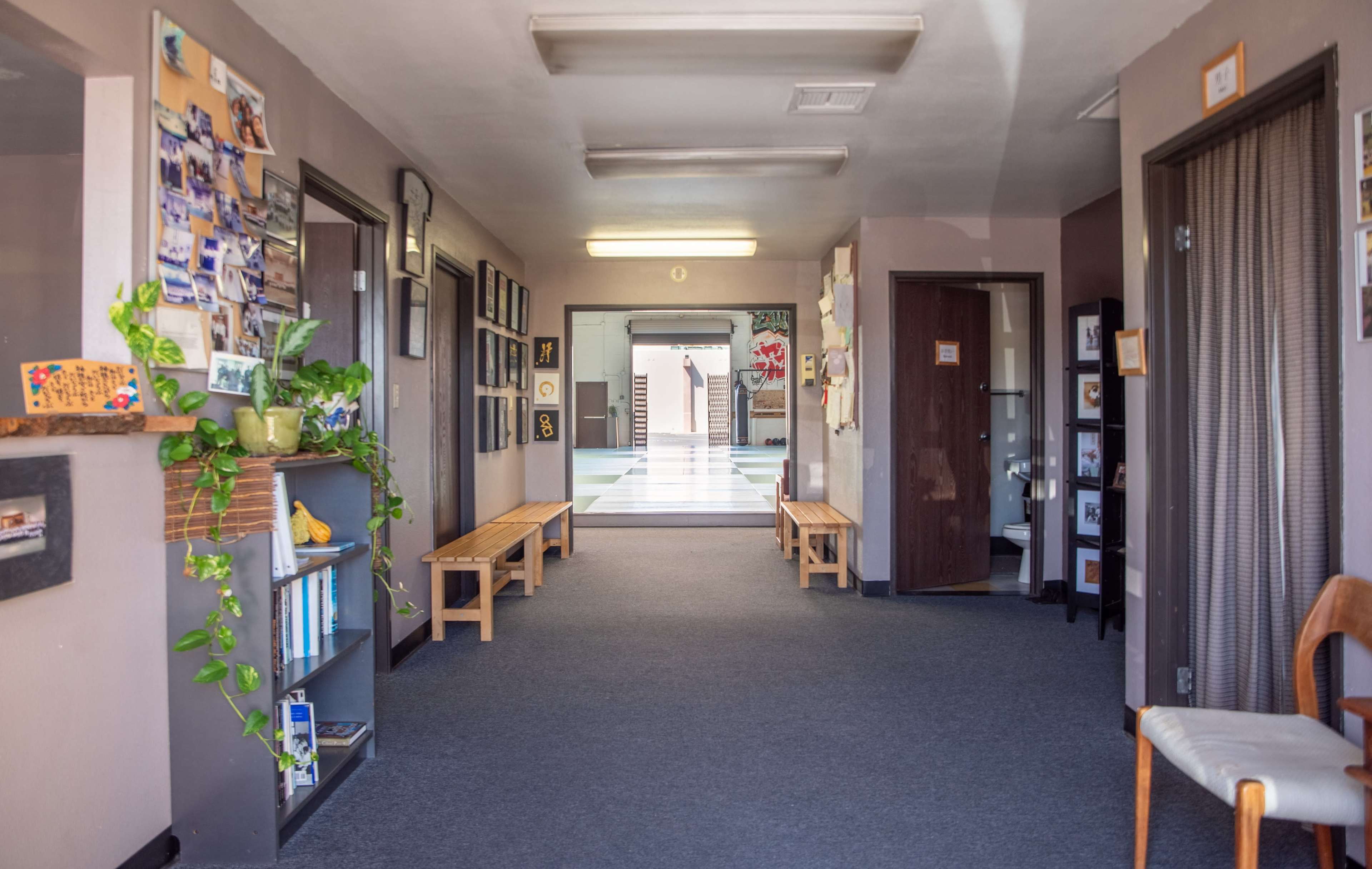 A hallway lined with benches and framed photographs, with doors on either side leading to rooms, and a view of an open area at the end.