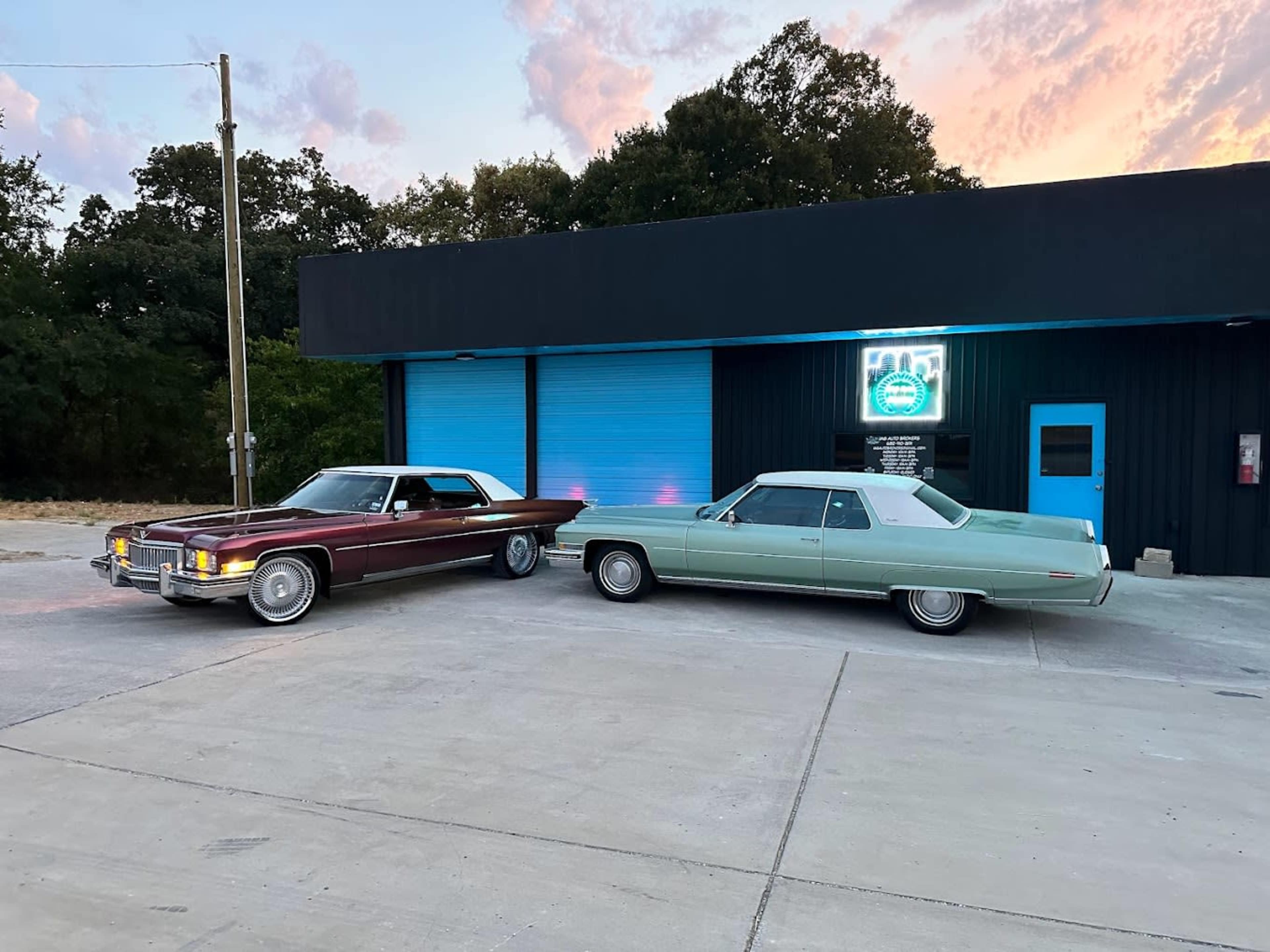 Two classic cars, a maroon coupe and a mint green sedan, are parked in front of a black building with a blue-lit sign at dusk.