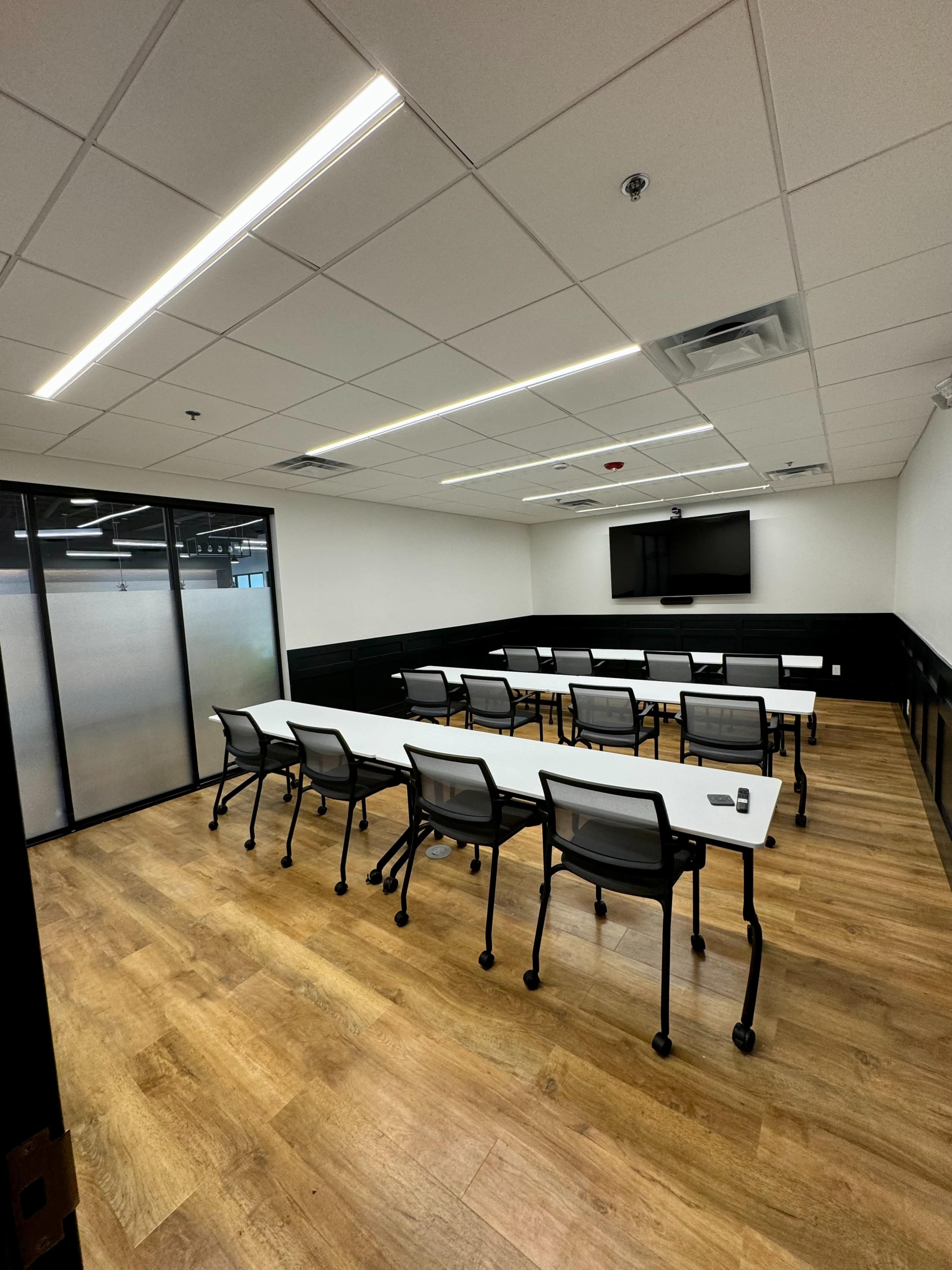 A modern conference room with rows of tables and chairs facing a flat-screen TV on the wall.