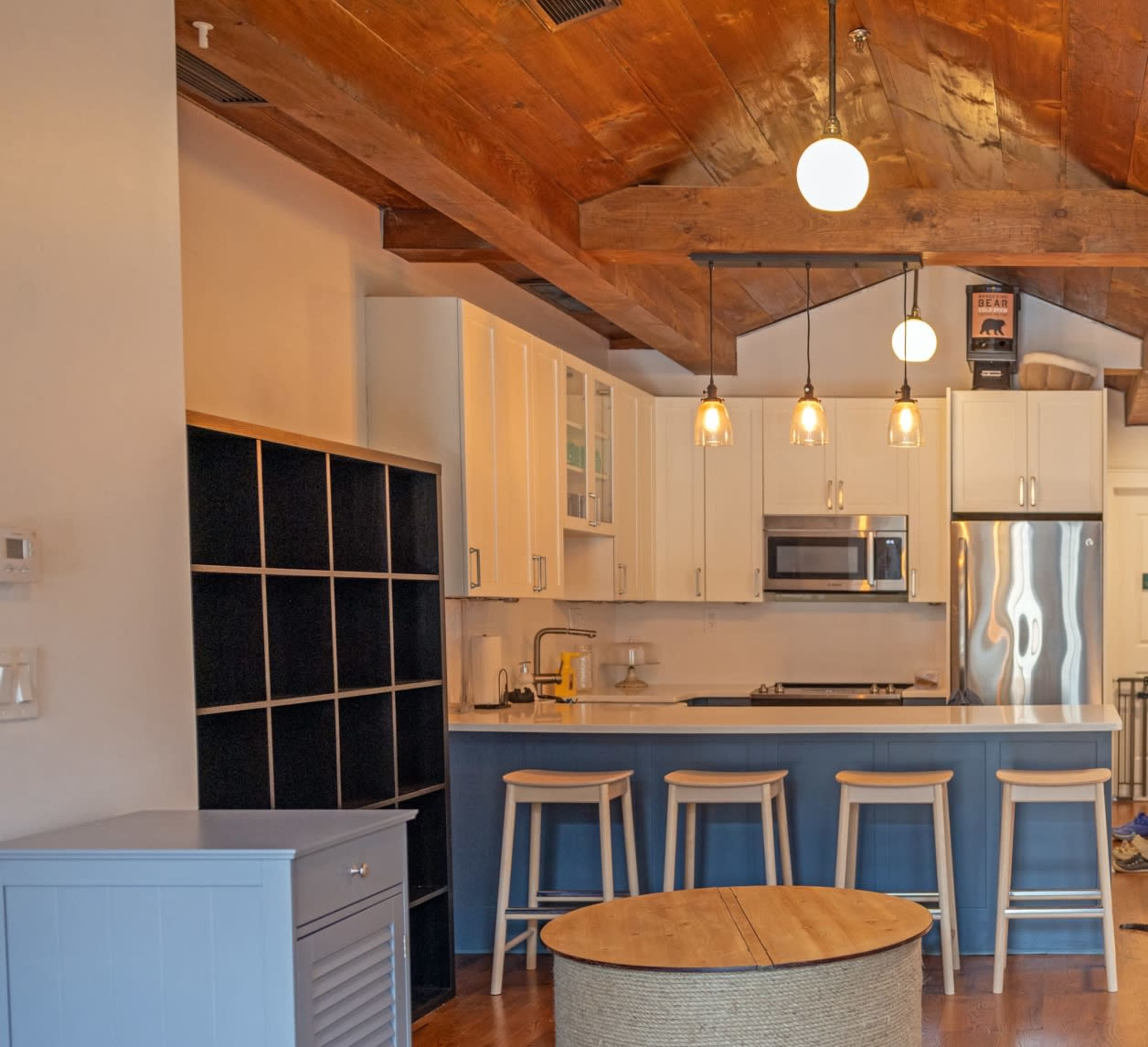 The image shows a modern kitchen with blue cabinetry, wooden beams overhead, and three bar stools at the counter.
