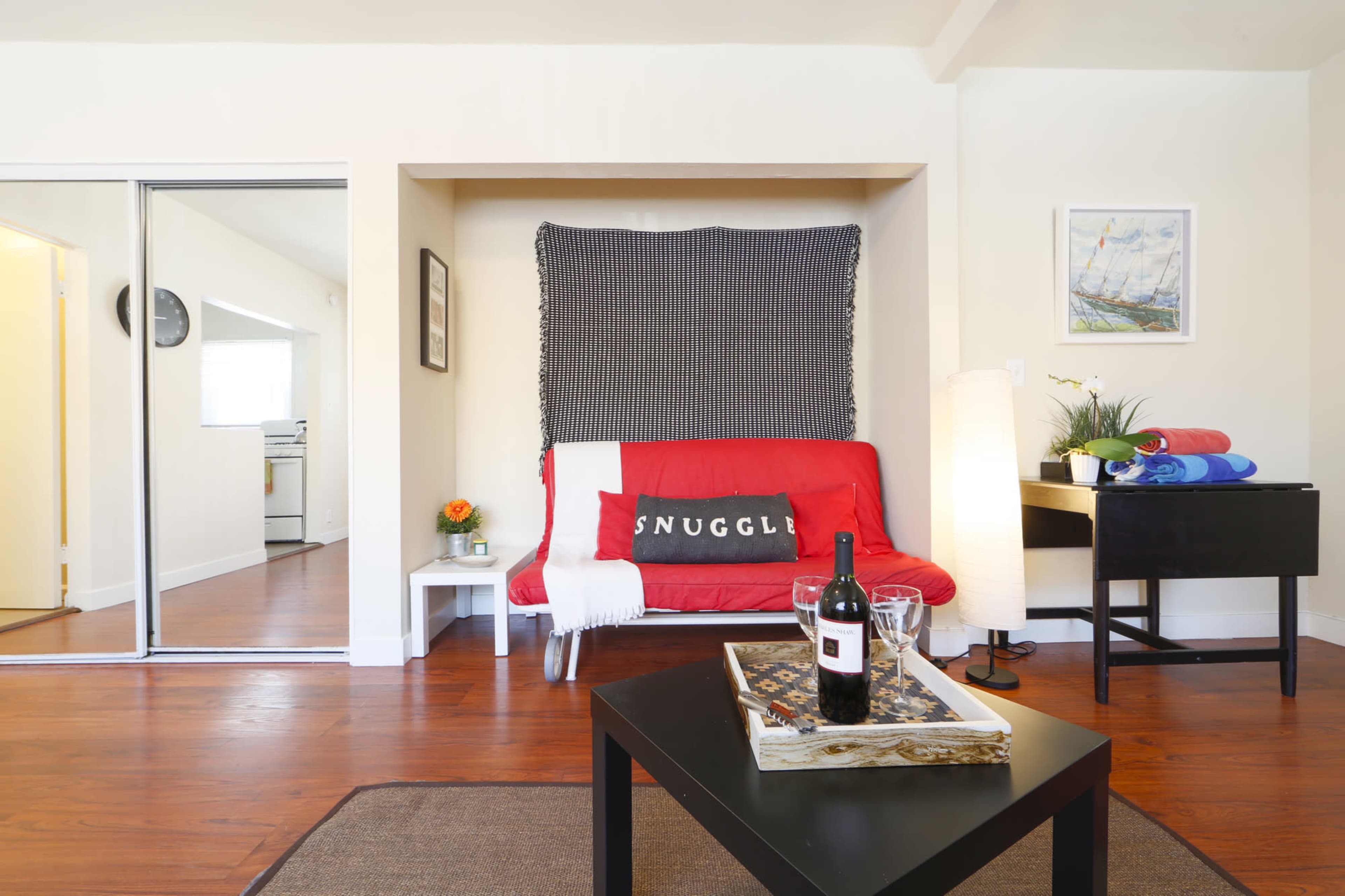 A cozy living space featuring a red futon with a "SNUGGLE" pillow, a small white table with a flower vase, and a black side table with a wine bottle and glasses.