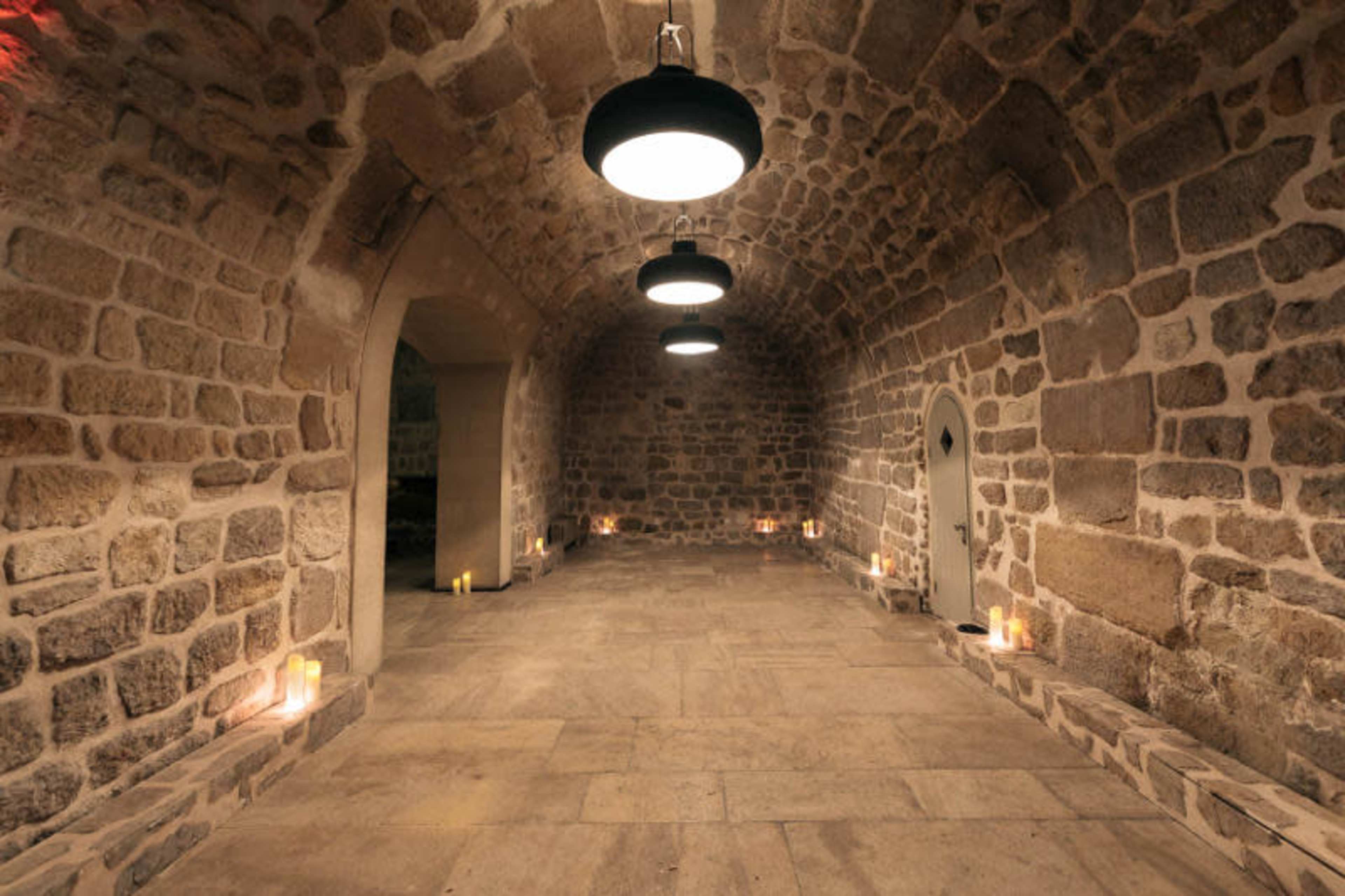 A stone-walled room with three hanging lights and candles placed along the perimeter of the floor.