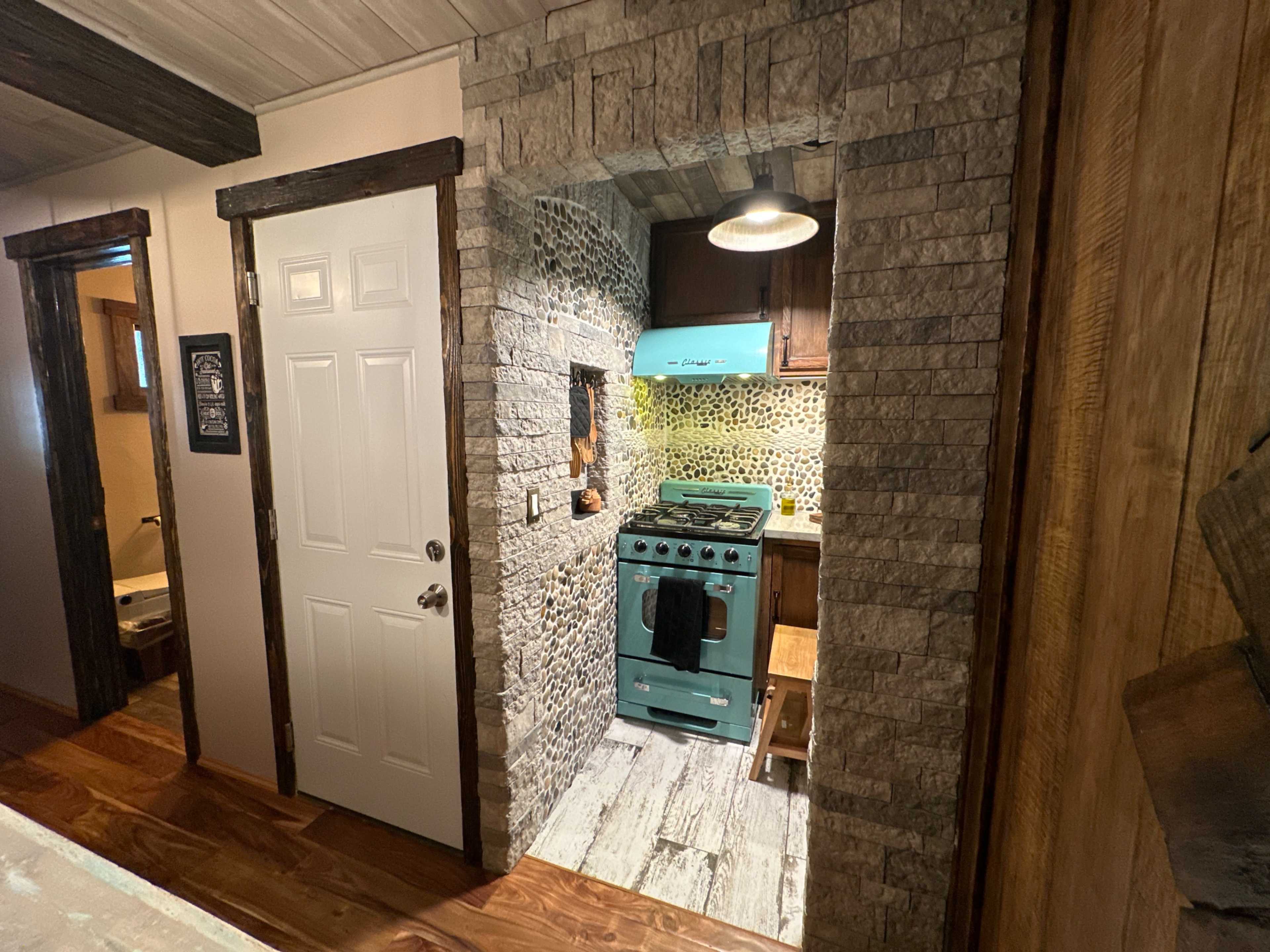 A small kitchen area with a retro teal stove, stone walls, and wooden accents, visible from a hallway.