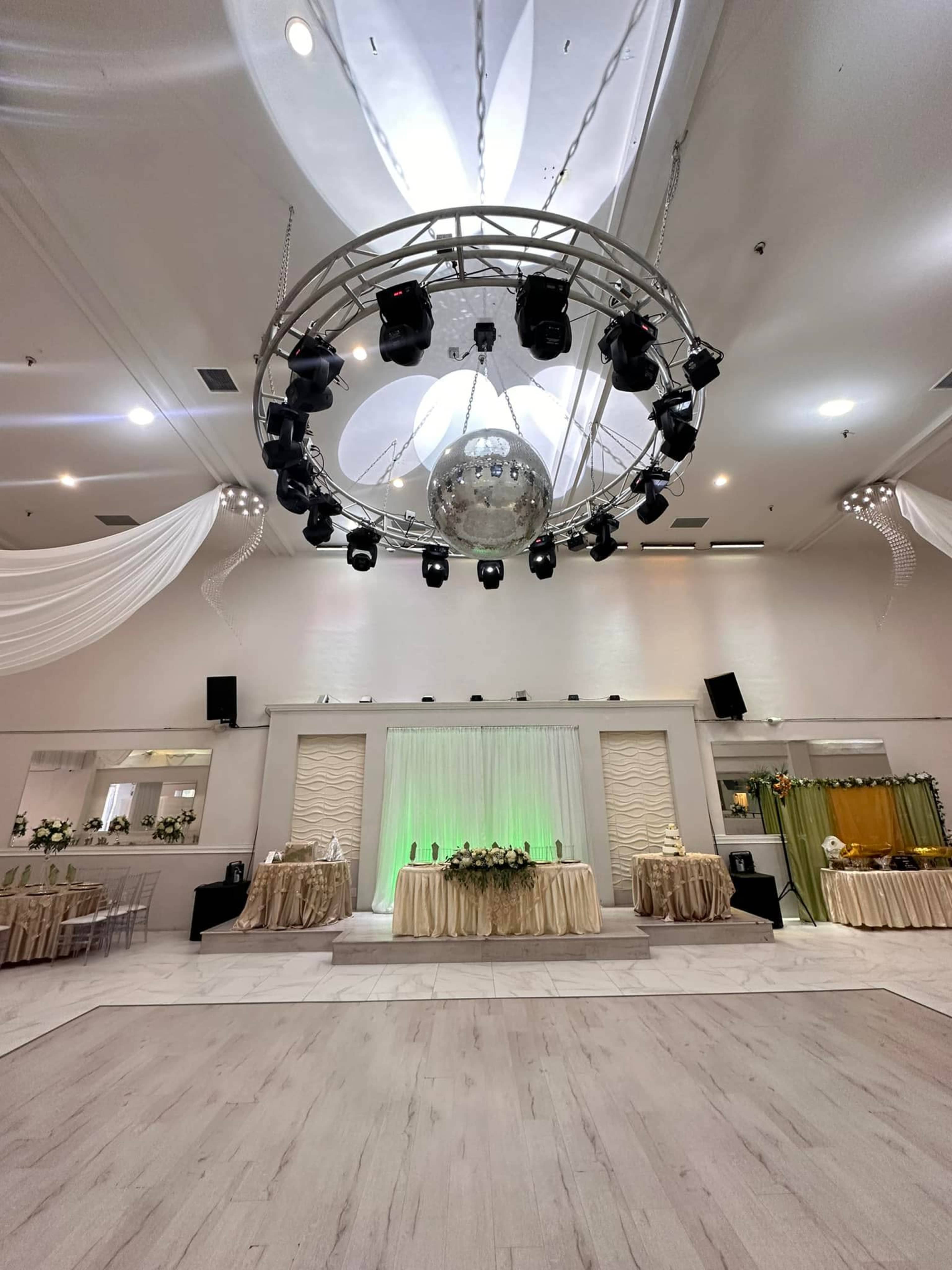The image shows a spacious event hall set up for a celebration, featuring a large disco ball suspended from the ceiling, with tables adorned in beige and green decorations.