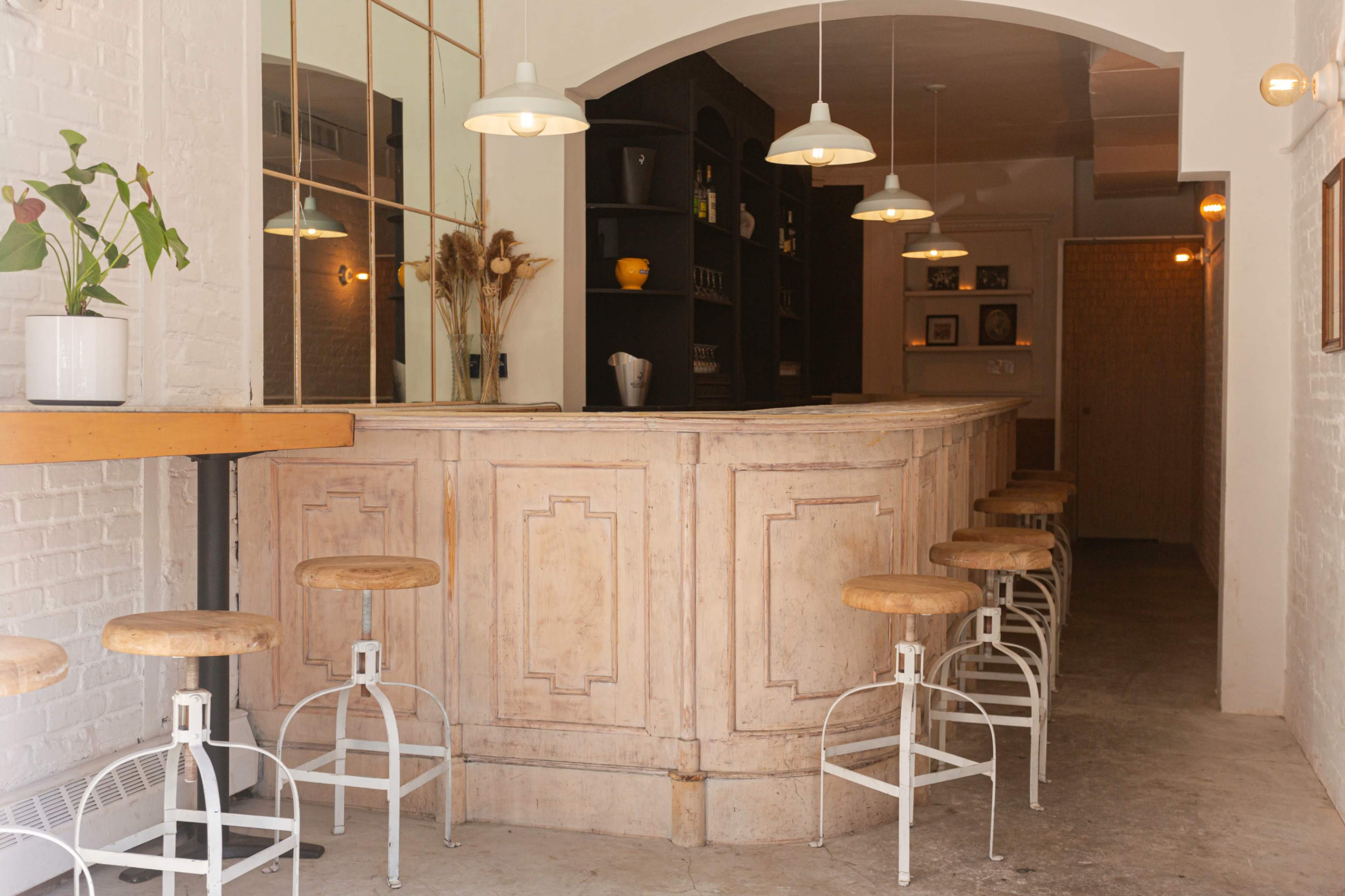 The image shows a minimalist bar area with a light wood counter and several high stools arranged neatly, illuminated by pendant lights.