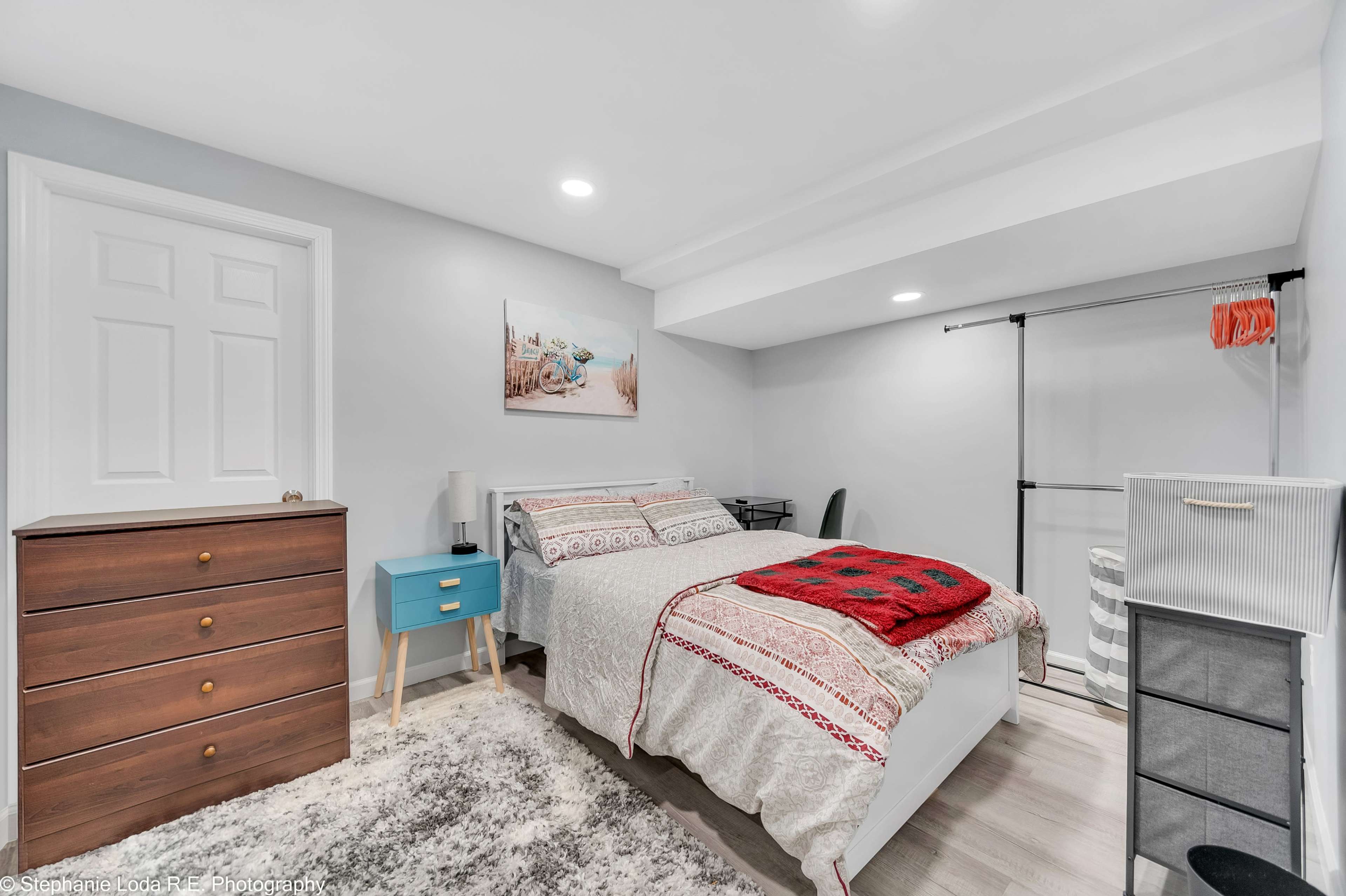 A small bedroom featuring a bed with a patterned duvet, a wooden dresser, a blue nightstand, and a gray and white striped storage container.
