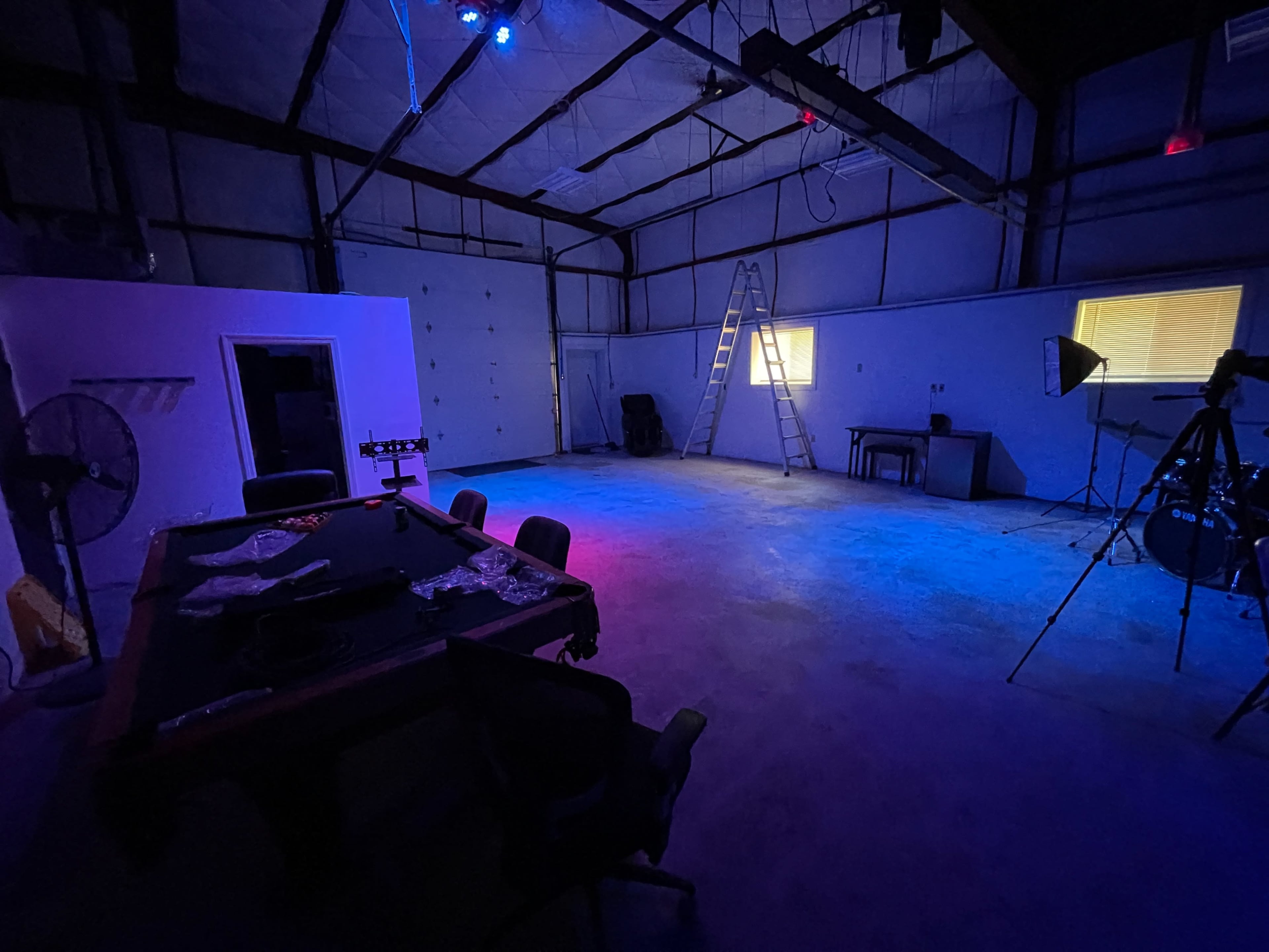 An empty industrial space features a table with chairs, a ladder against the wall, and various lighting equipment illuminated in purple and blue.