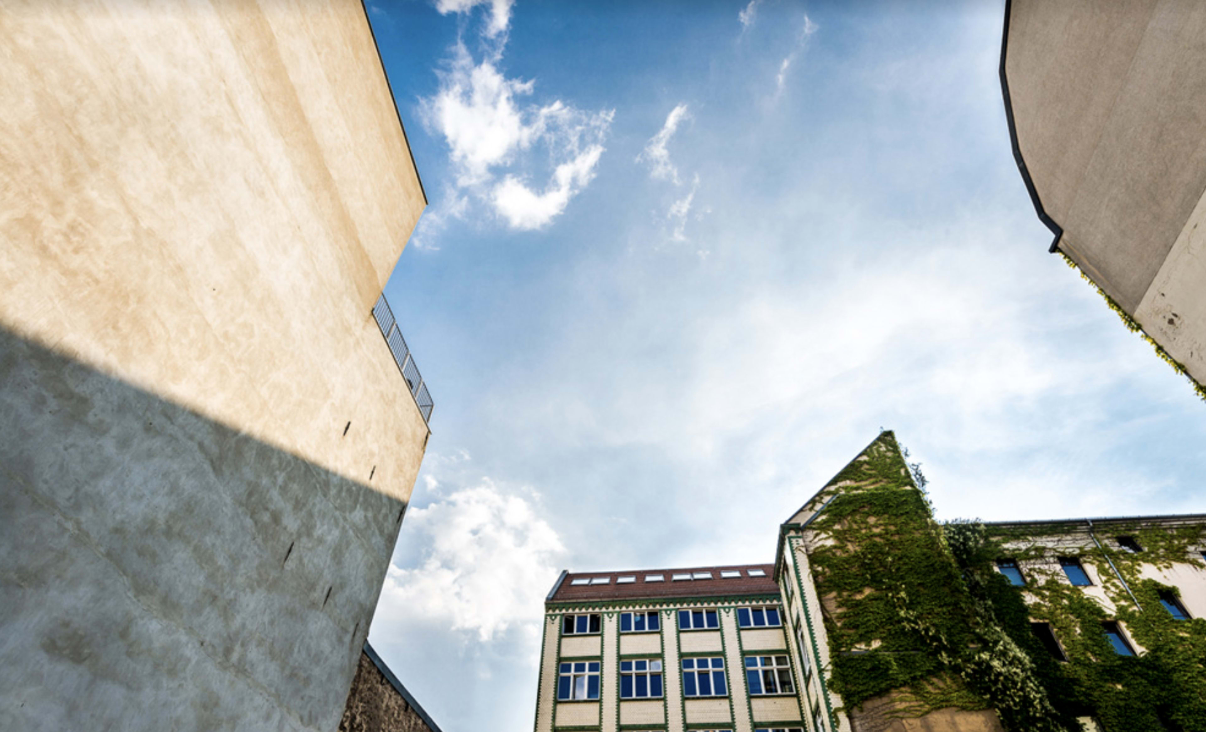 The image shows a view of buildings from a narrow alley, with a bright blue sky and scattered clouds above.
