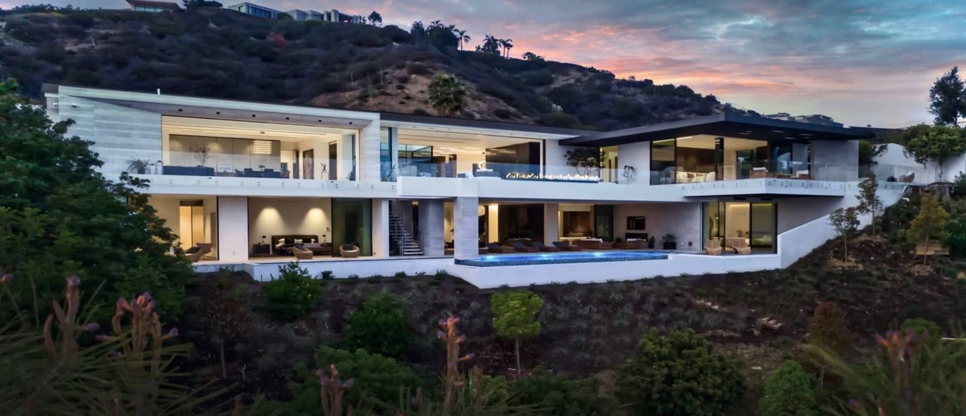 A modern hillside mansion features multiple levels, large glass windows, and an outdoor pool surrounded by lush greenery.