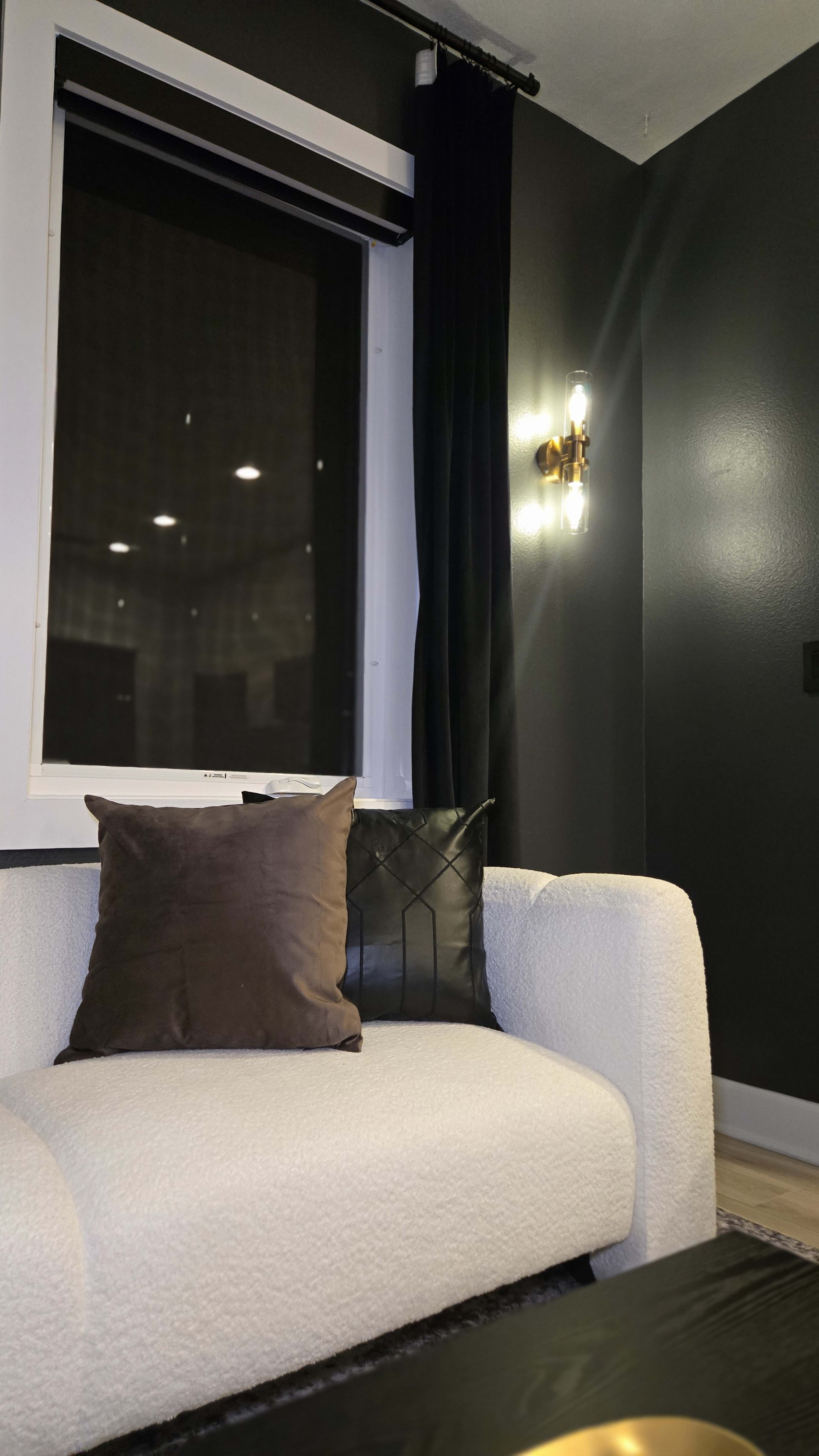 A cozy corner of a room features a plush white sofa with a brown cushion, near a window with dark curtains and wall-mounted lighting.