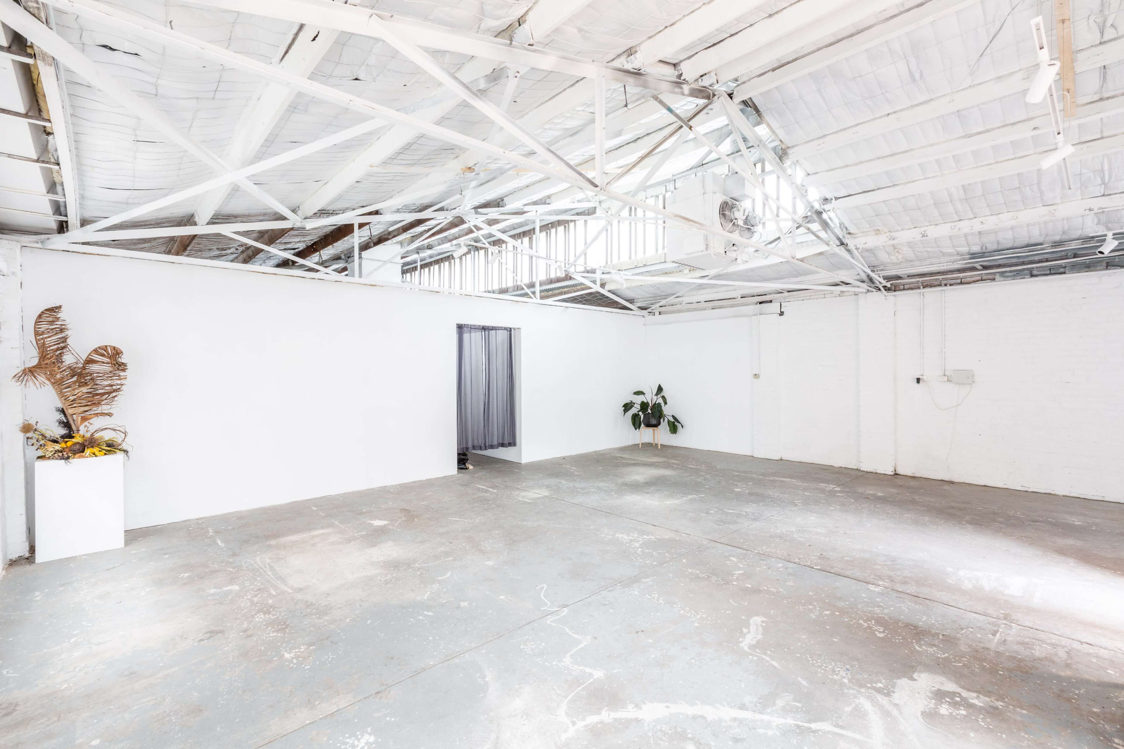 A spacious, empty interior of a warehouse with white walls, exposed beams, and a concrete floor, featuring a plant and a decorative element in one corner.