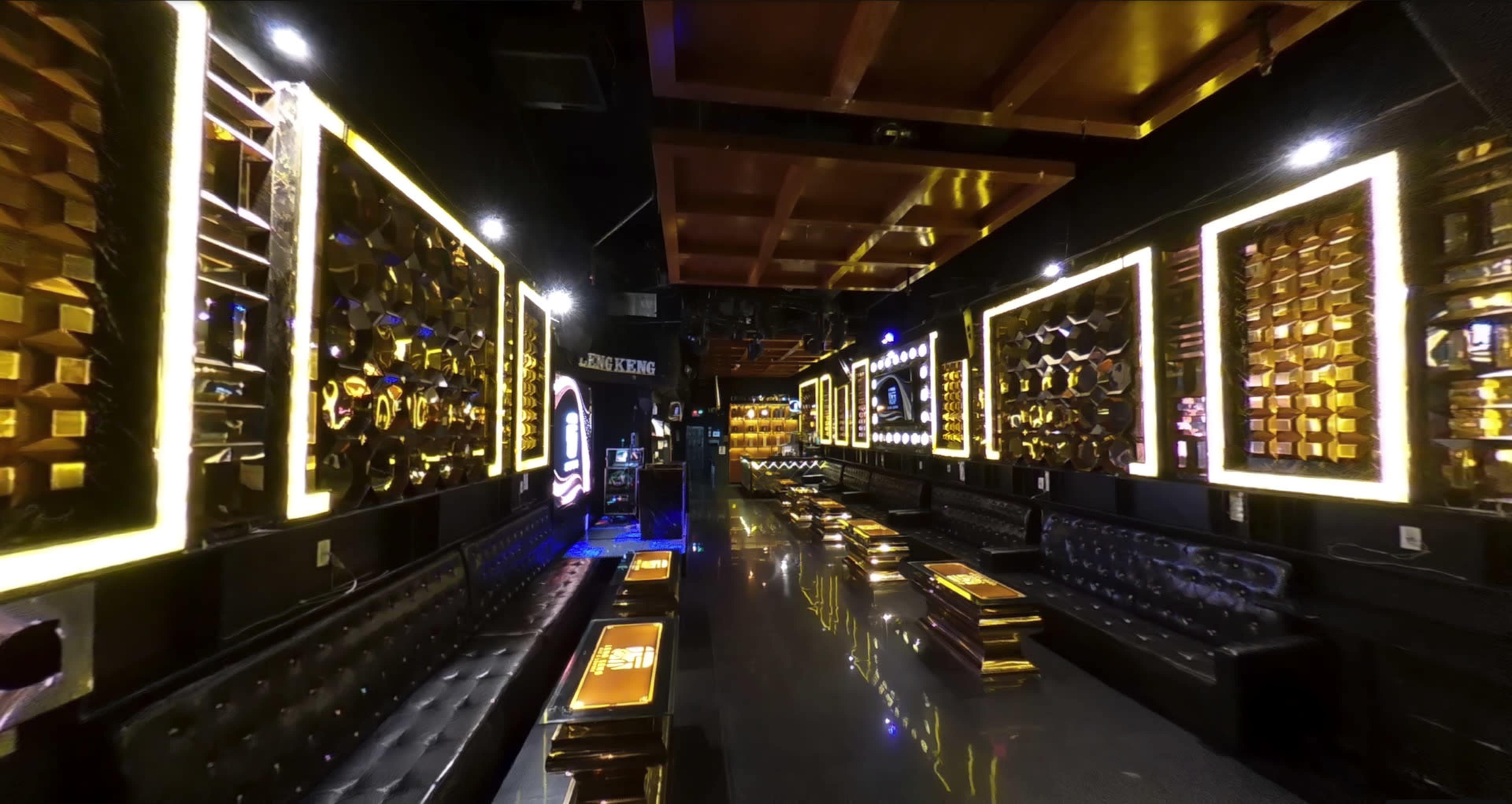 The interior of a dimly lit lounge features black leather seating, illuminated gold-framed wall designs, and sleek reflective surfaces.