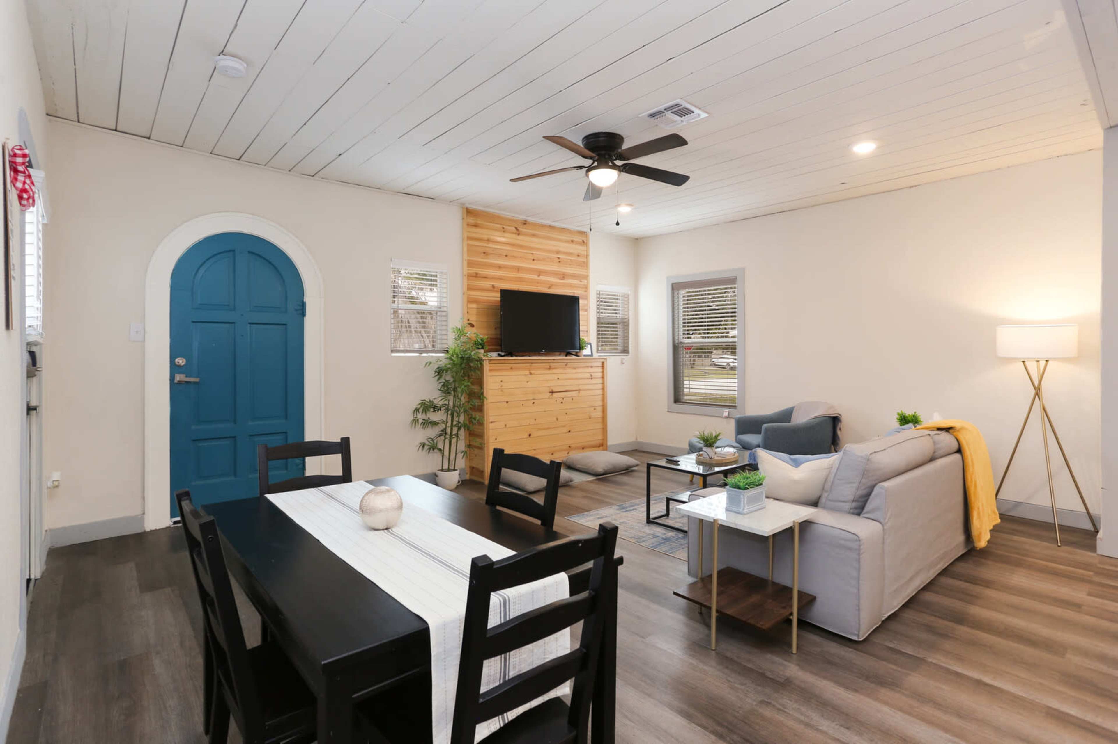 The image shows a modern living space with an open layout featuring a dining table, a sofa, a wooden accent wall with a mounted television, and a blue door.