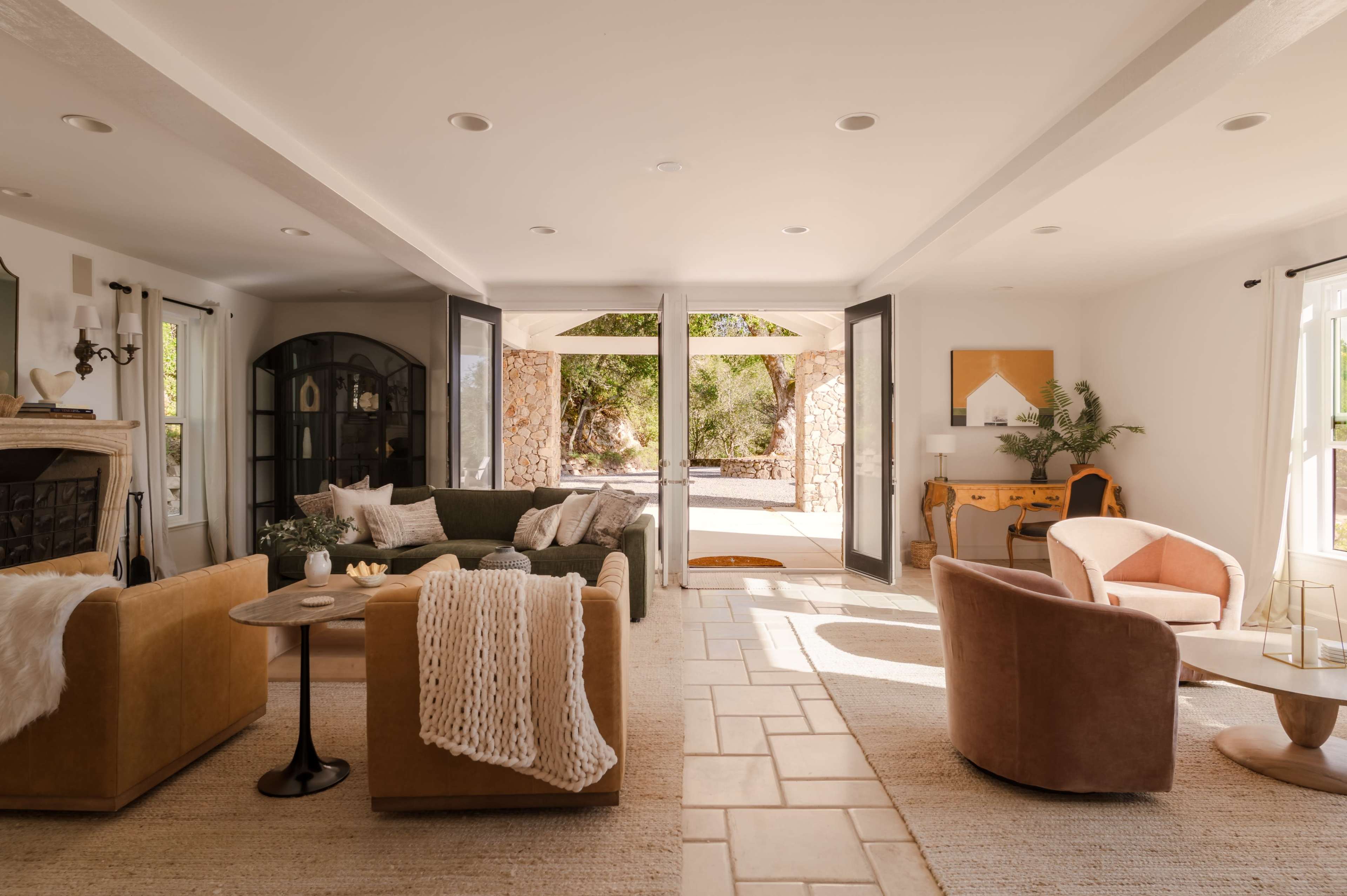 The image shows a modern living room with open glass doors leading to an outdoor space, featuring a mix of comfortable seating arrangements and decorative elements.