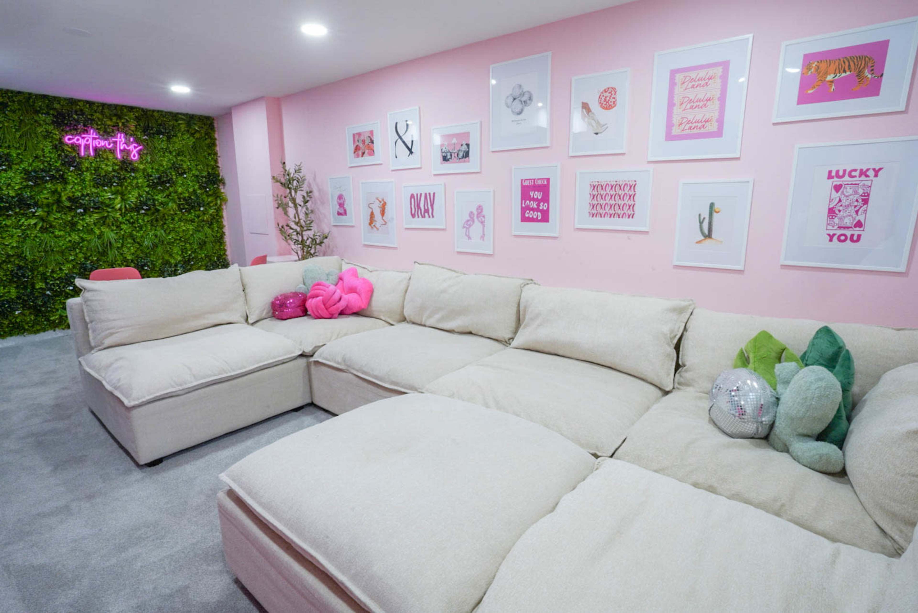 A cozy lounge area featuring a large sectional sofa, pink walls adorned with framed artworks, and a green plant wall in the background.