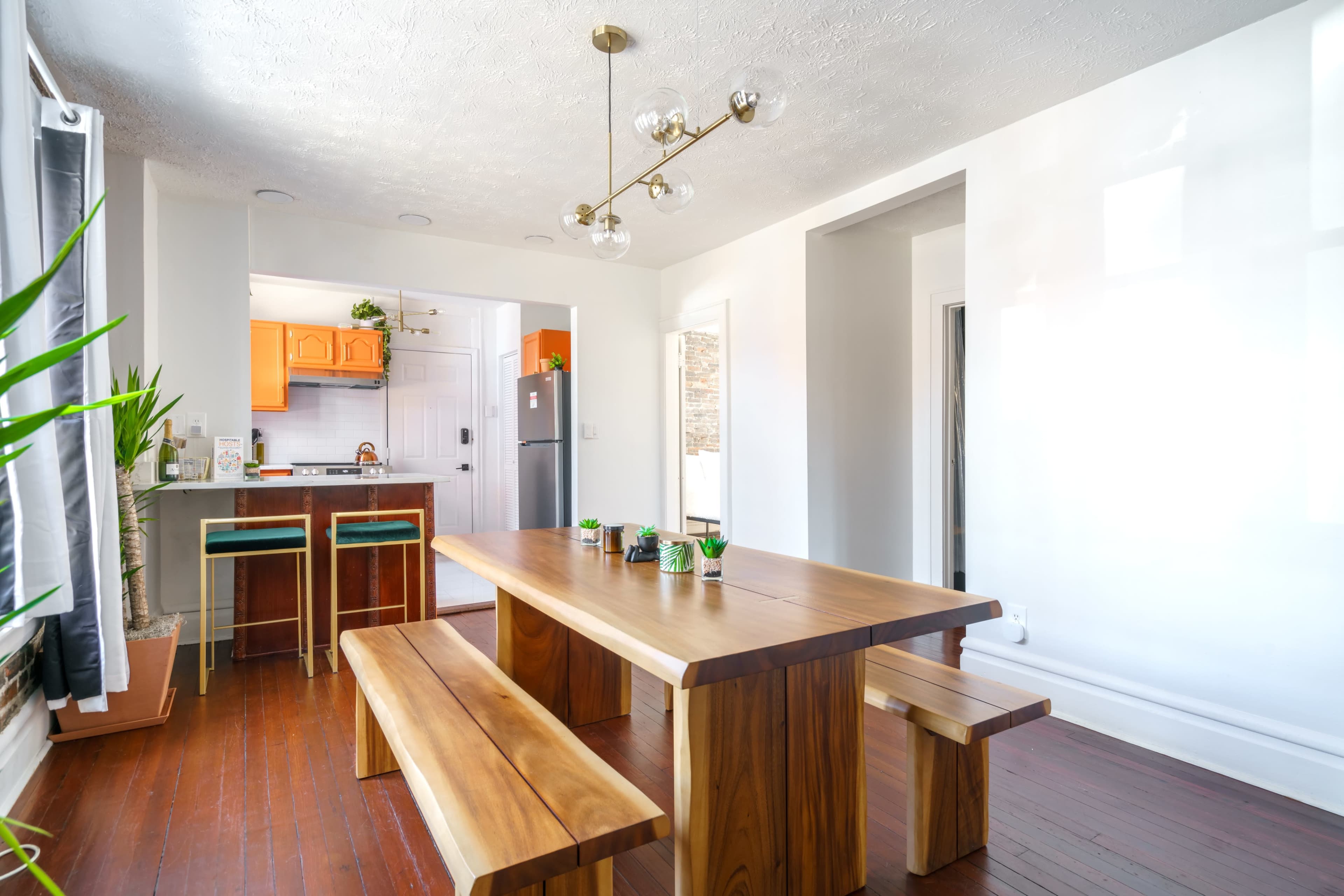 The image shows a well-lit dining area featuring a large wooden table with benches, surrounded by plants, leading to a kitchen with orange cabinets in the background.